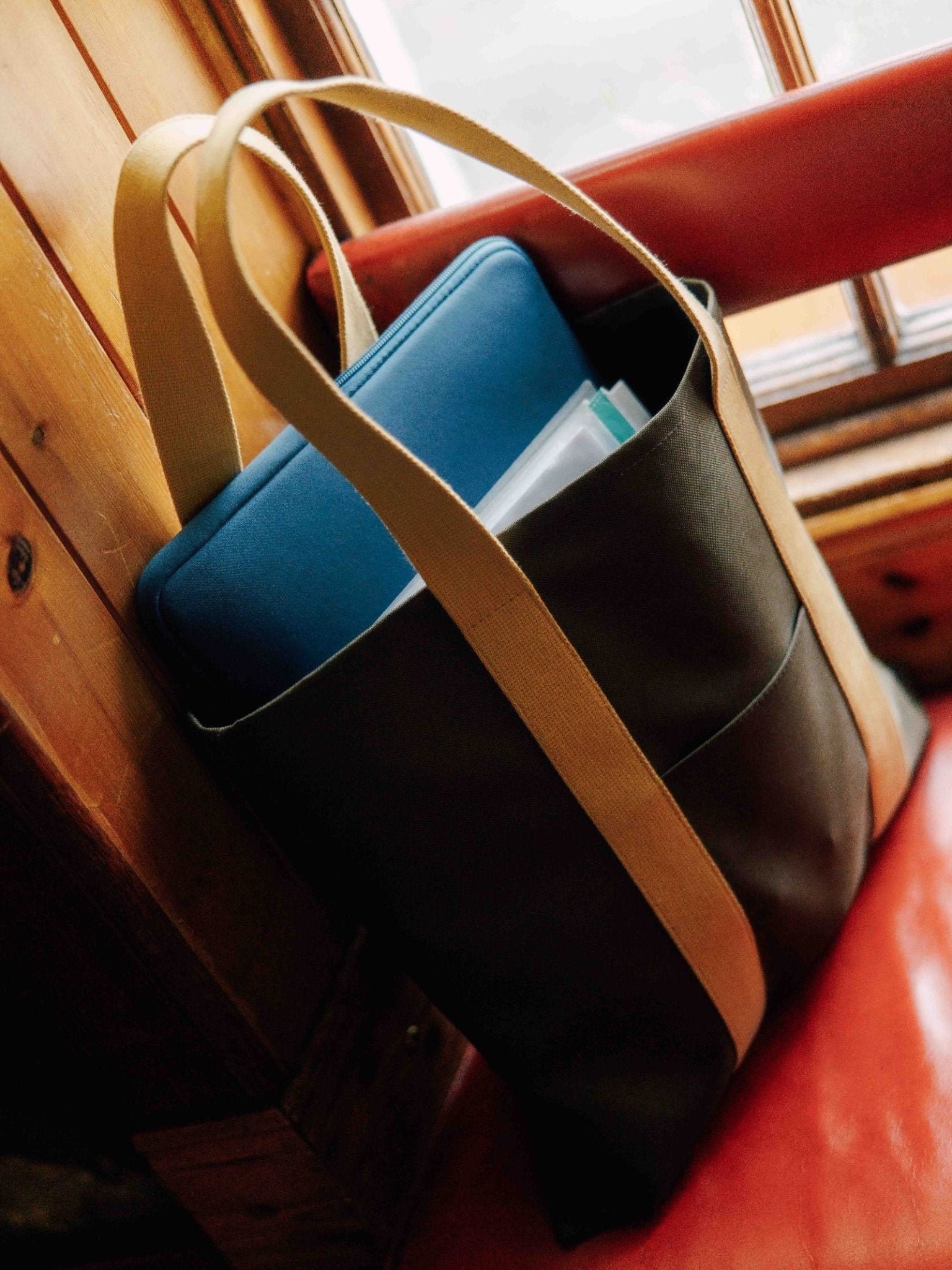 A Campbell's of Beauly Utility Packaway Tote with tan straps sits on a red cushioned seat by a wooden wall. A blue laptop sleeve and papers peek from this lightweight, durable black canvas bag.