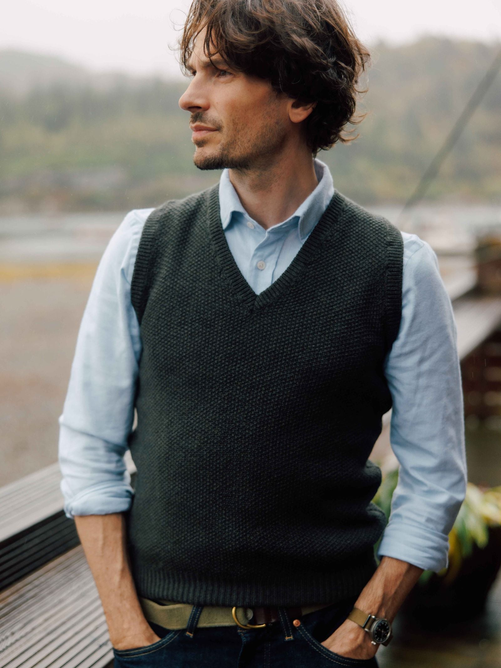 A man with wavy brown hair stands outdoors, looking to the side. He wears Campbell's of Beauly's Auld Stock Twill Shirt in light blue and a dark grey knitted vest, hands in his jeans pockets against a softly blurred natural background.