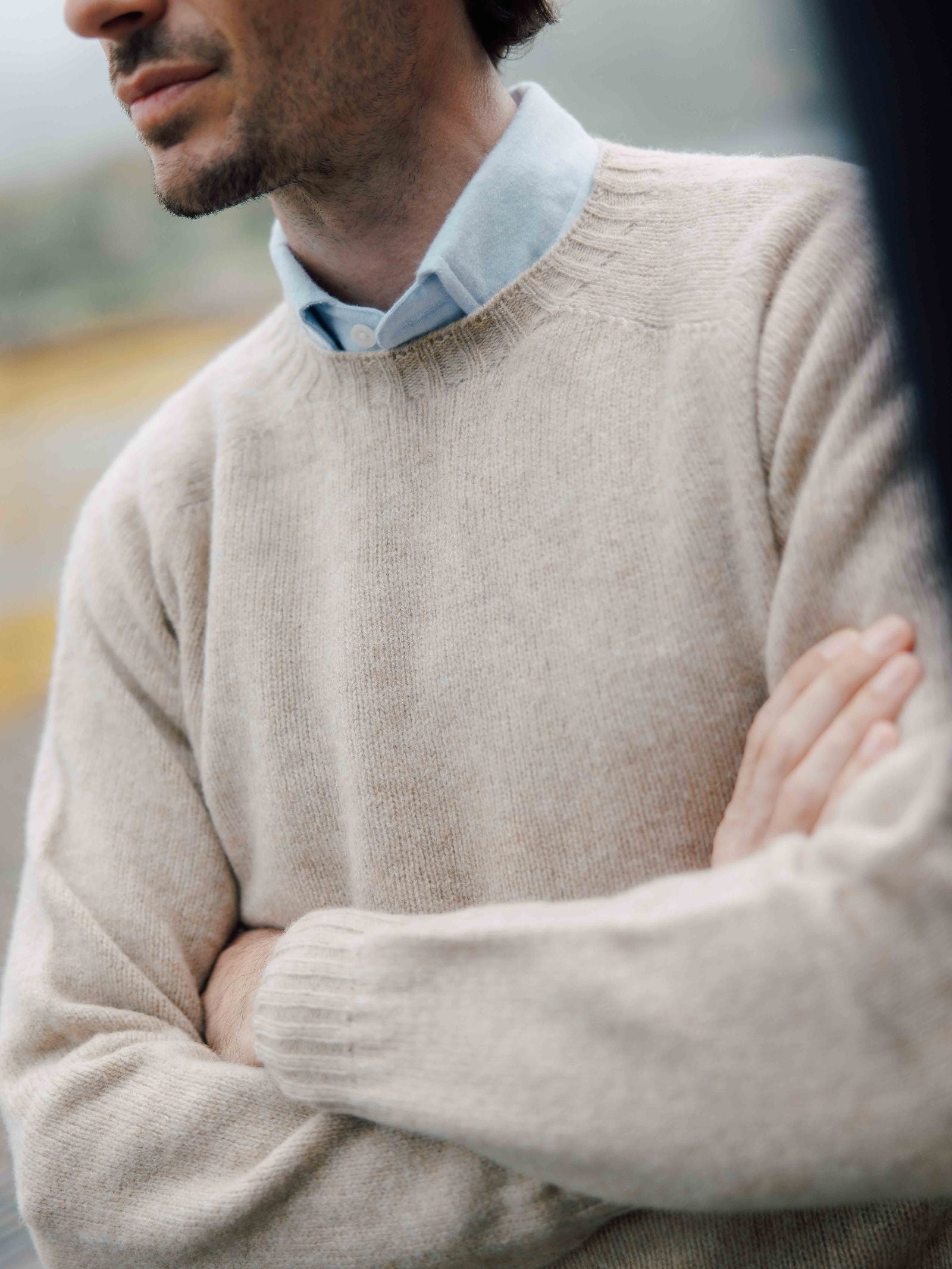 A man with a light beard wears the beige Shetland Jumper by Campbell's of Beauly over a blue collared shirt, standing with arms crossed against a softly blurred background, showcasing classic Scottish knitwear style.