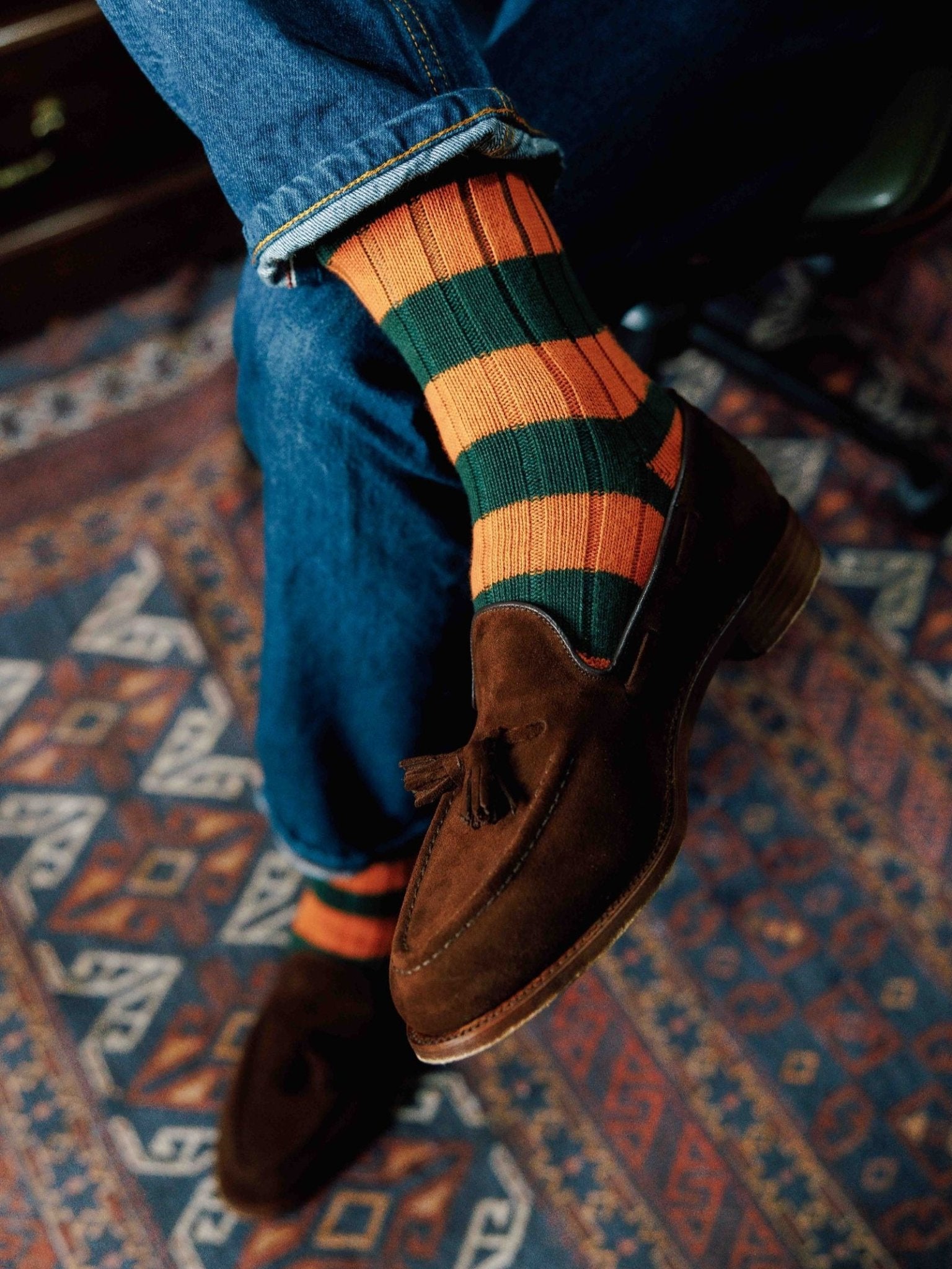 A person wearing blue jeans, brown suede tassel loafers, and Campbell's of Beauly Stripe Socks sits cross-legged on a patterned rug.