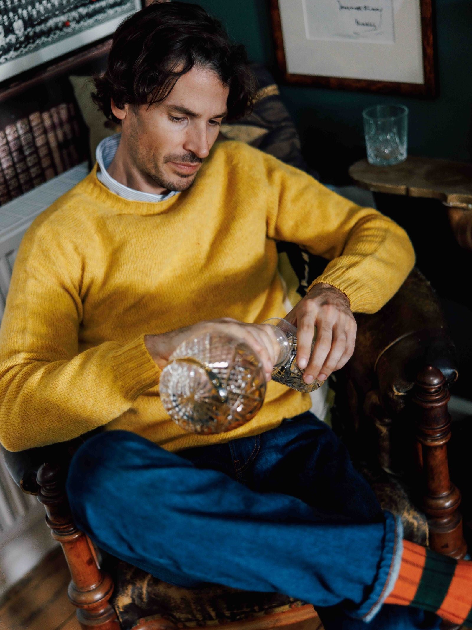 A man with wavy brown hair, dressed in a cozy yellow Campbell's of Beauly Shetland Jumper and blue jeans, sits relaxed in a chair, pouring a drink from a glass decanter with one leg crossed in a warm indoor setting.