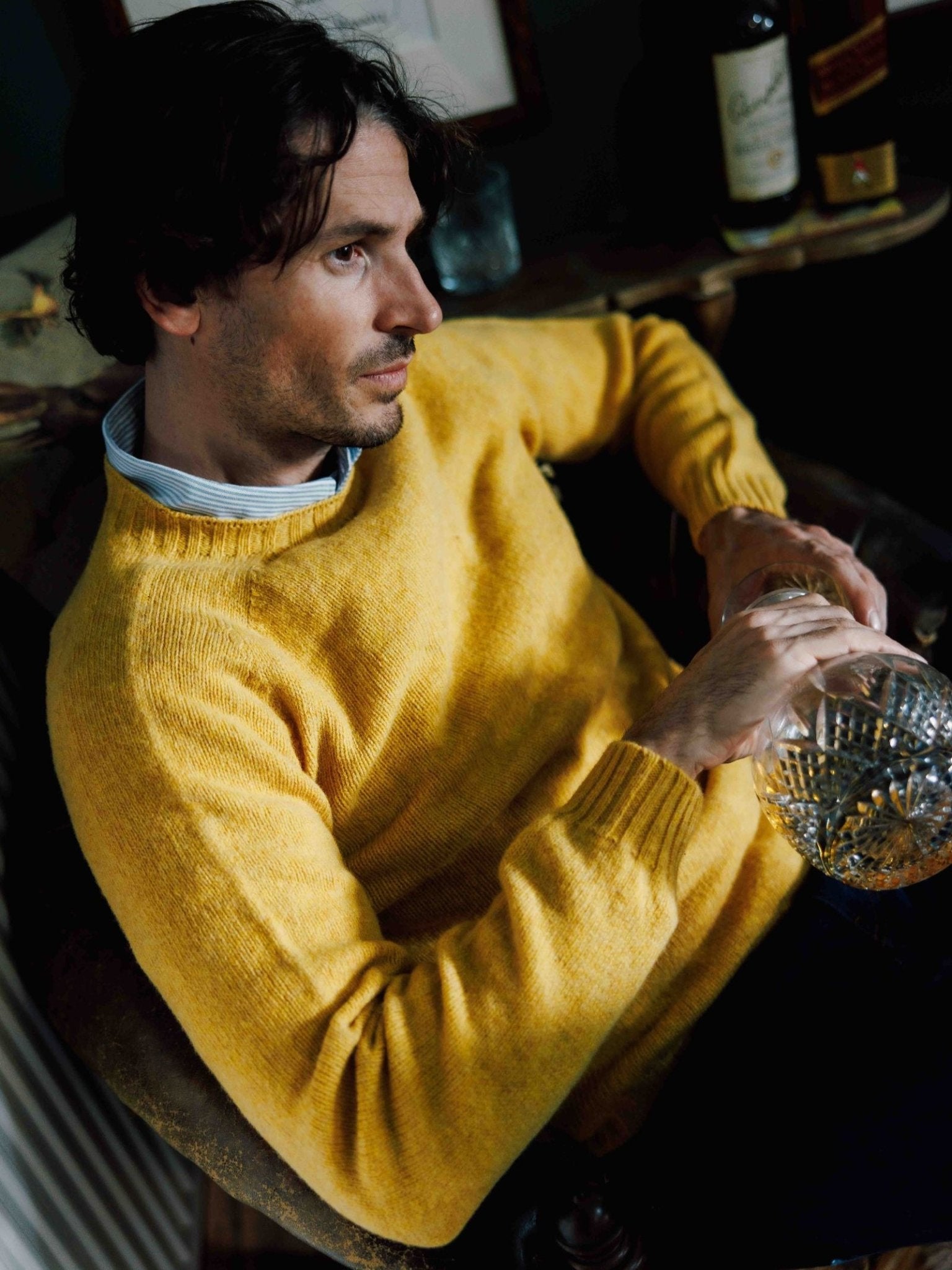A man with wavy brown hair wears a yellow Campbell's of Beauly Shetland Jumper over a collared shirt, sitting thoughtfully in a chair and holding a glass decanter, with bottles visible on the table behind him.