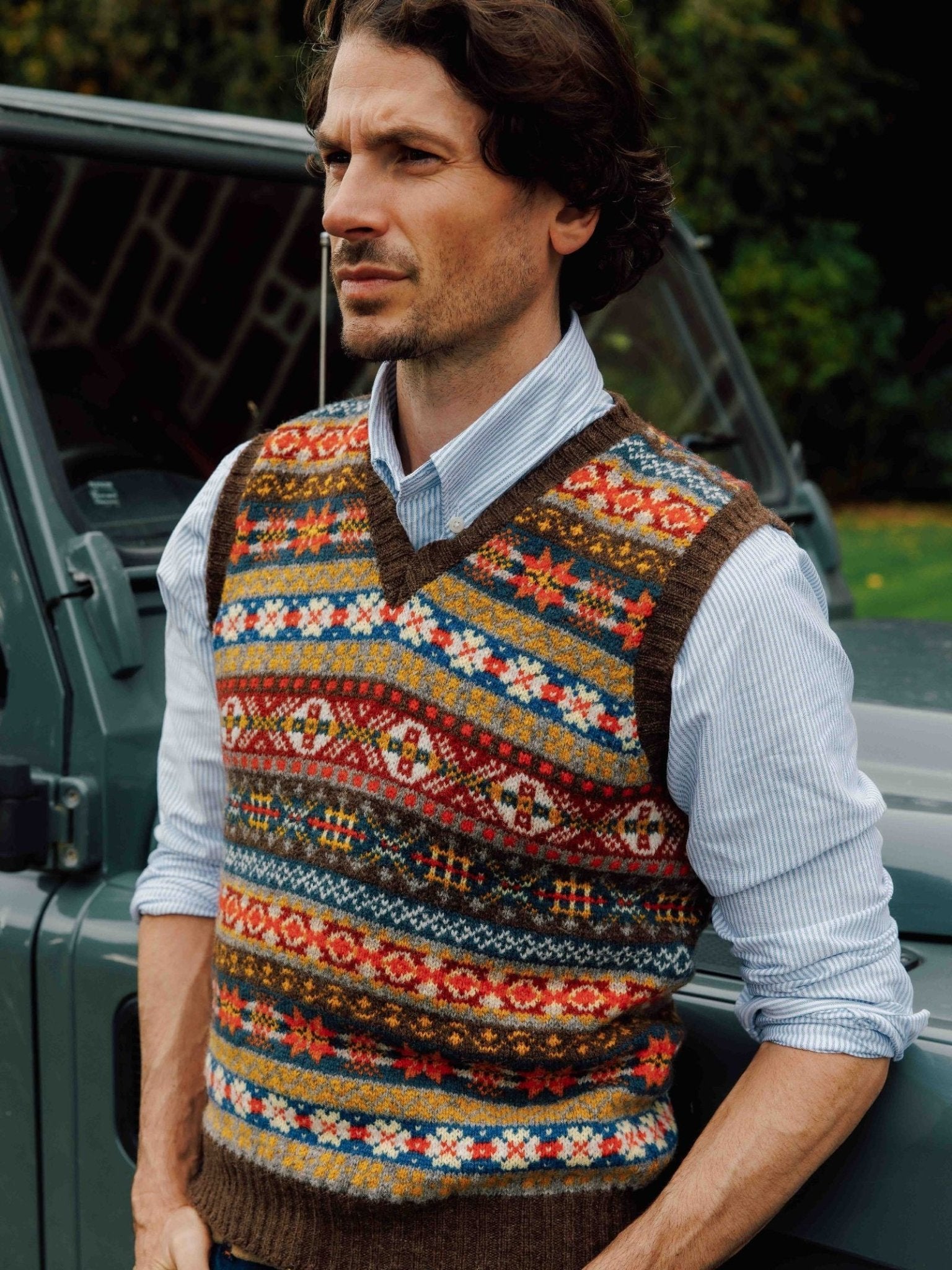 A man with wavy brown hair wears the Campbell's of Beauly Fairisle Slipover, a Shetland wool knit vest, layered over a striped button-up shirt while standing outdoors in front of a gray vehicle.
