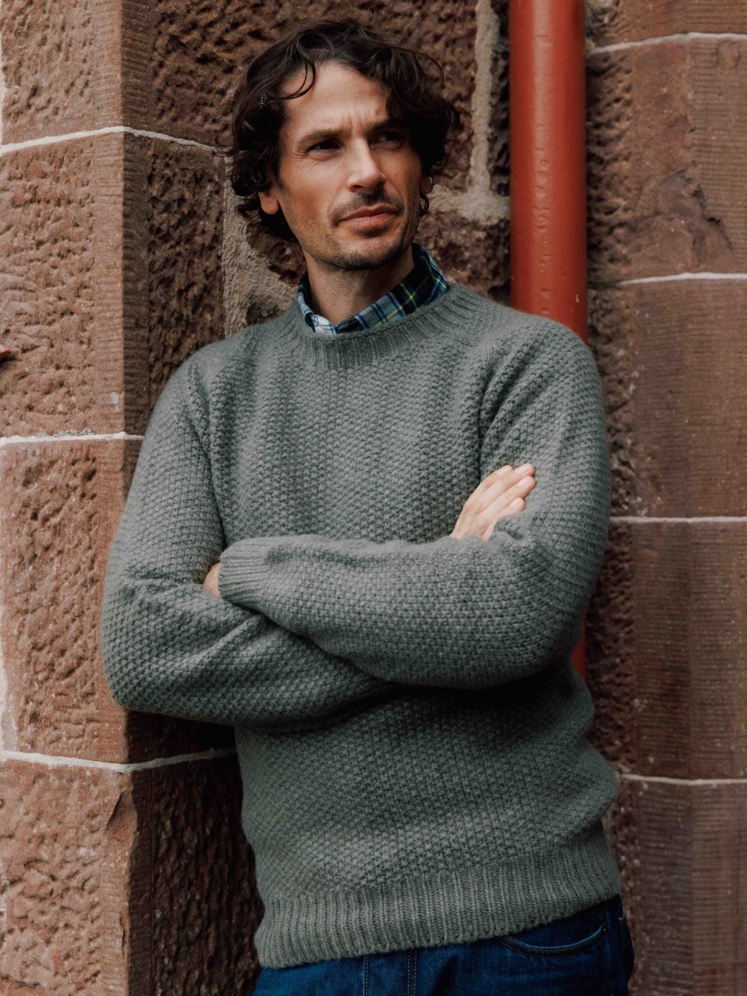 A man with curly brown hair and a beard stands by a stone wall, wearing the Campbell's of Beauly Moss Stitch Jumper—a classic crew neck style knitted in Scotland—over a plaid shirt, arms crossed, gazing slightly to the side.