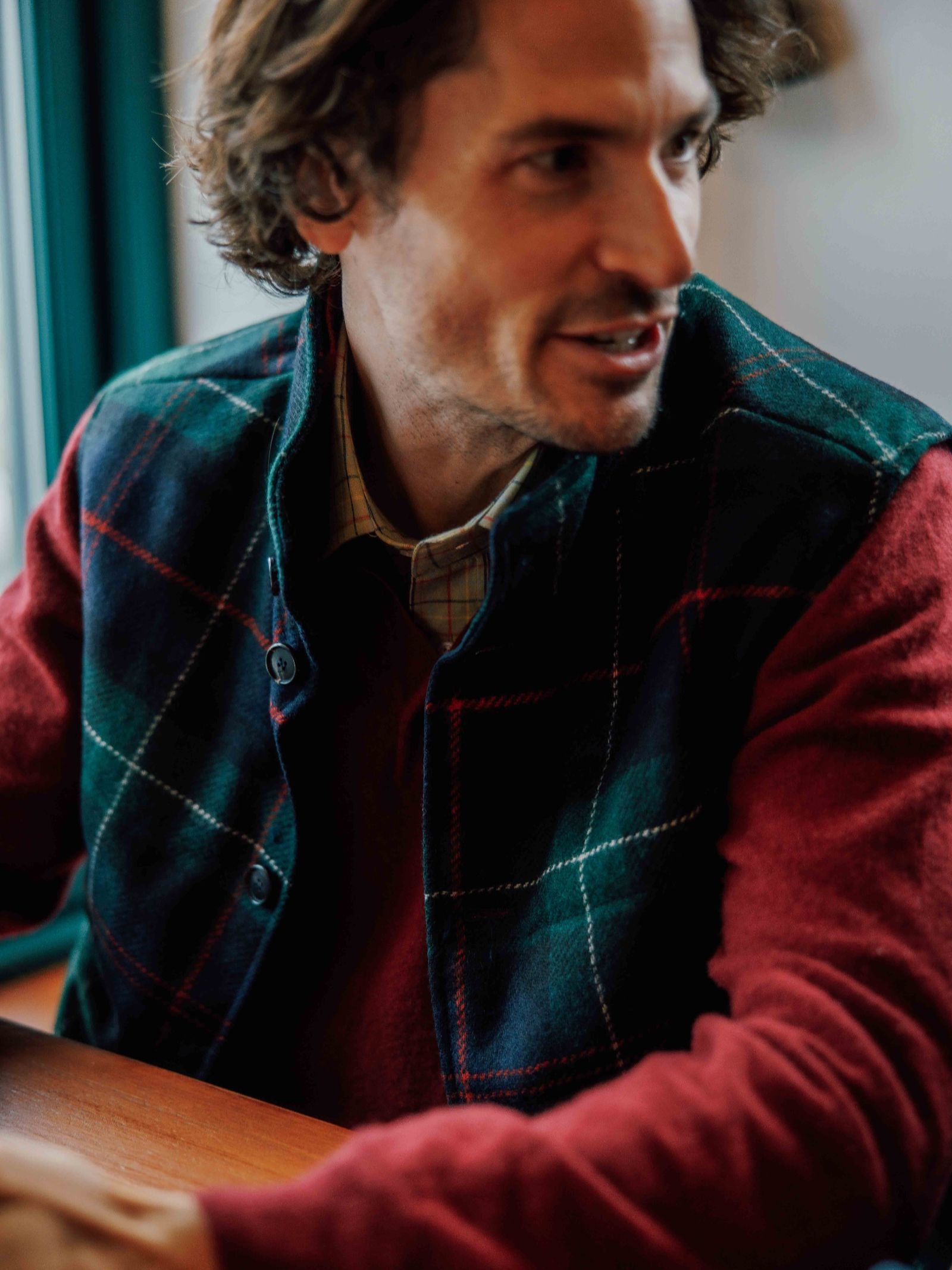 A man with wavy brown hair sits indoors, smiling slightly as he wears a red sweater layered with the Campbell's of Beauly Quilted Tweed Buttoned Gilet, his arm resting on a wooden table.