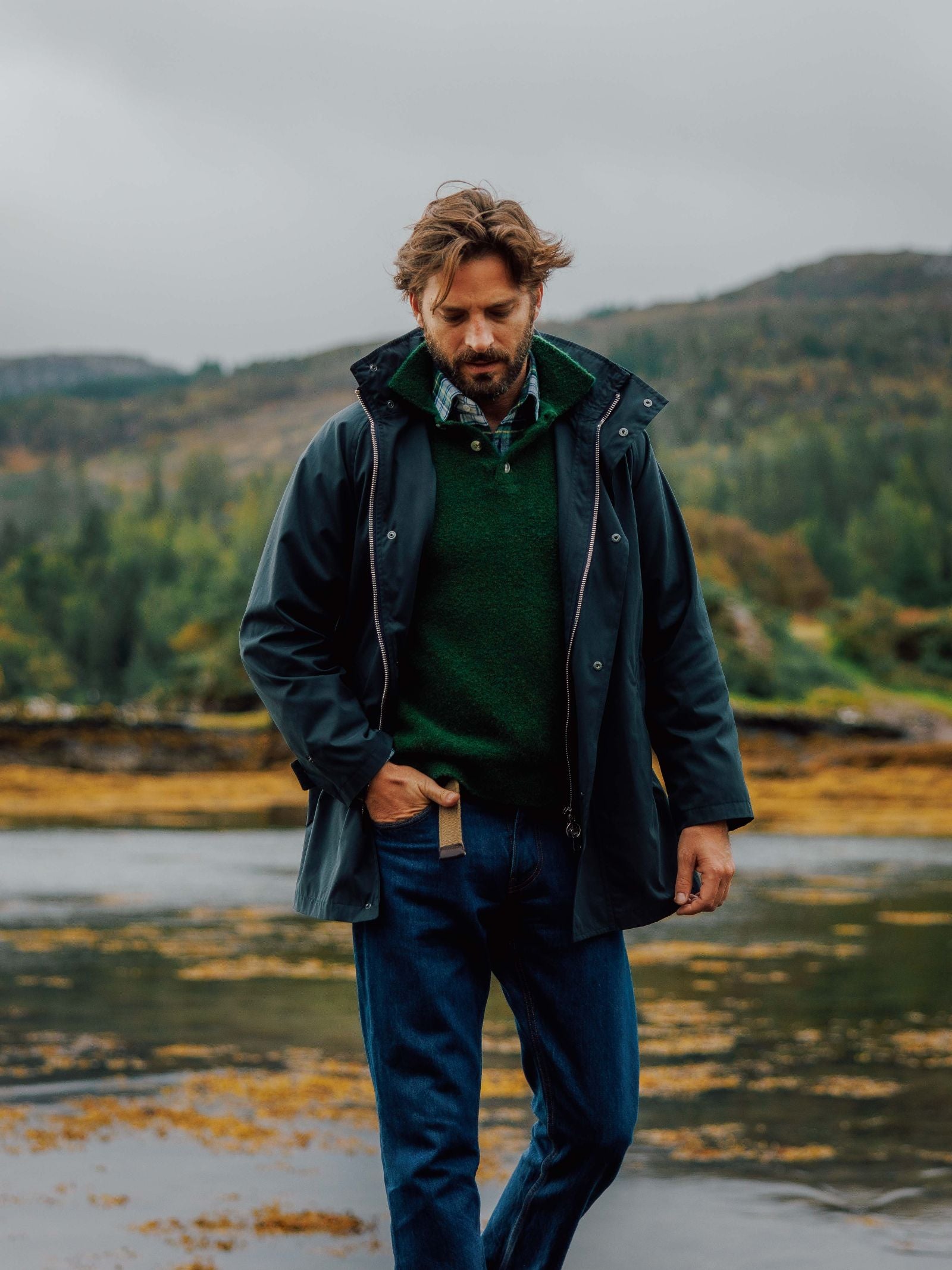 A man with brown hair and a beard walks outdoors by the water, wearing a dark raincoat, plaid shirt, jeans, and the Campbell's of Beauly Bouclé Polo Sweater. Hills, trees, and cloudy skies appear in the background.