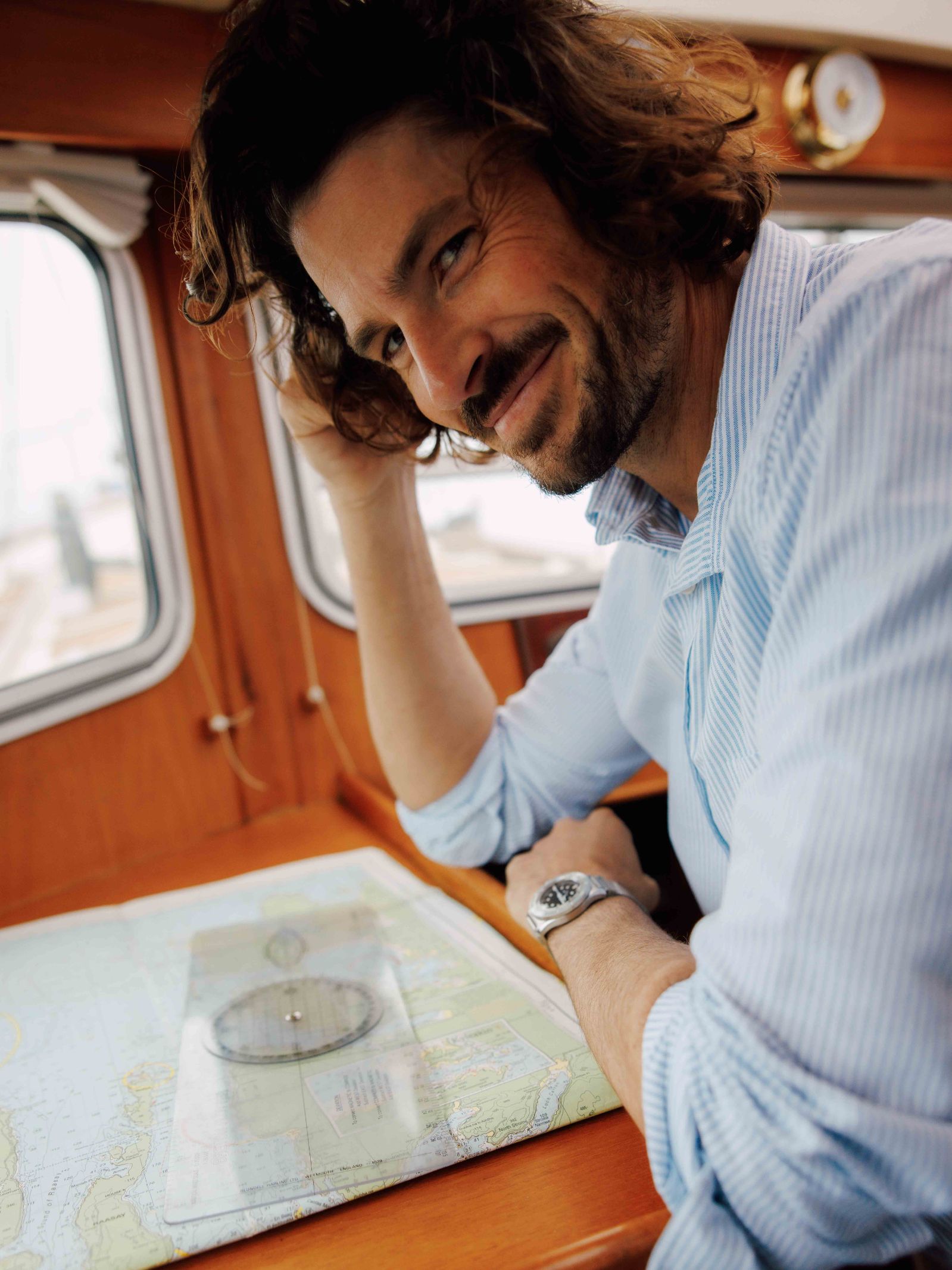 A man with wavy brown hair and a beard, wearing a light blue Campbells of Beauly Twill Shirt and a watch, sits at a wooden table on a boat, smiling slightly at the camera with his arm resting on a nautical map topped with a navigation tool.