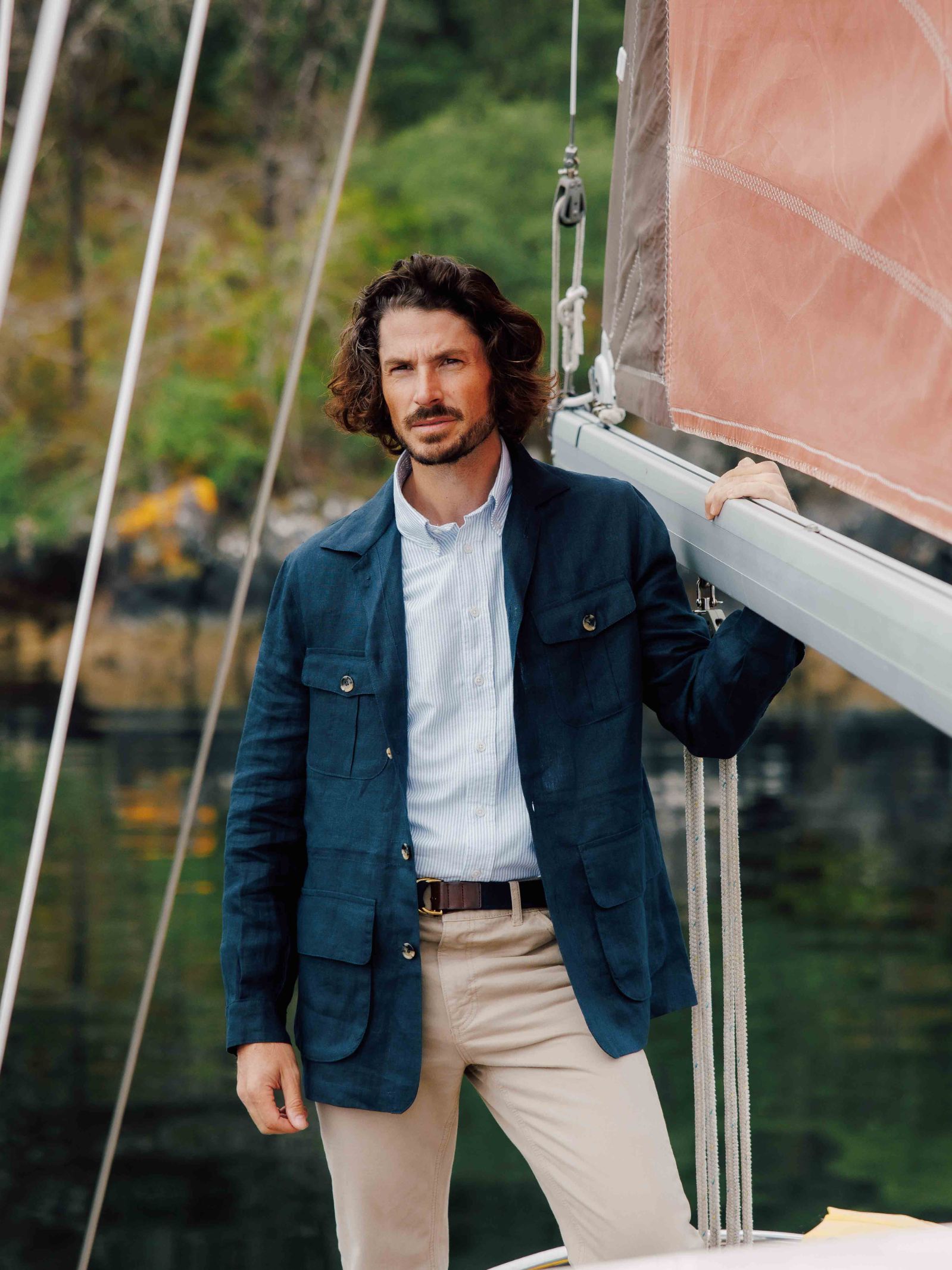 A man with wavy brown hair and a beard stands on a sailboat, wearing the Campbells of Beauly Linen Safari Jacket as a casual layer, one hand resting on the rigging. Green foliage and water form the background.