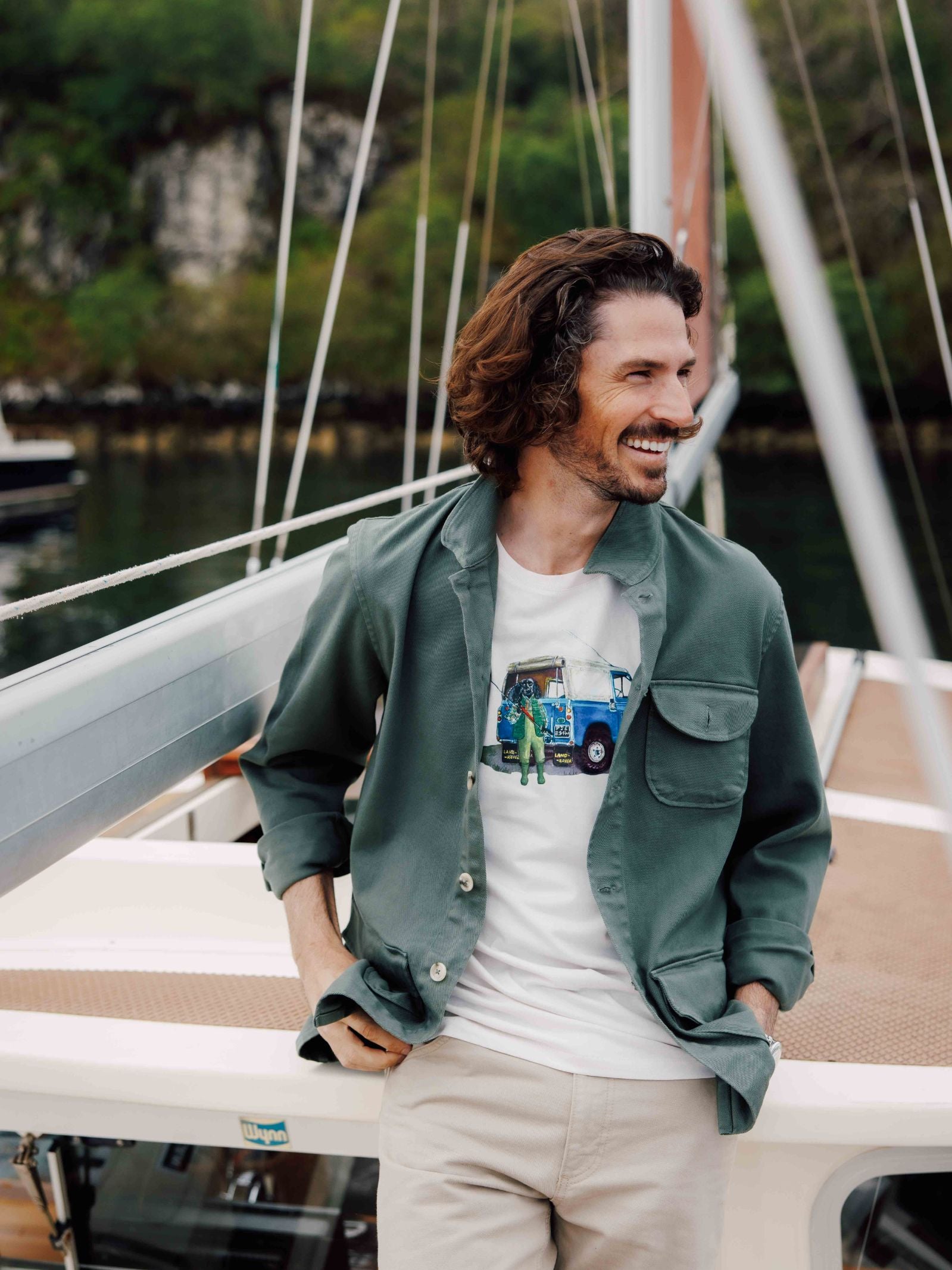 A smiling man with wavy brown hair and a beard stands on a boat, wearing a green jacket over the Land Rover Cocker T-Shirt by Campbells of Beauly and beige pants. The backdrop features water, greenery, and part of the boat’s mast and railing.
