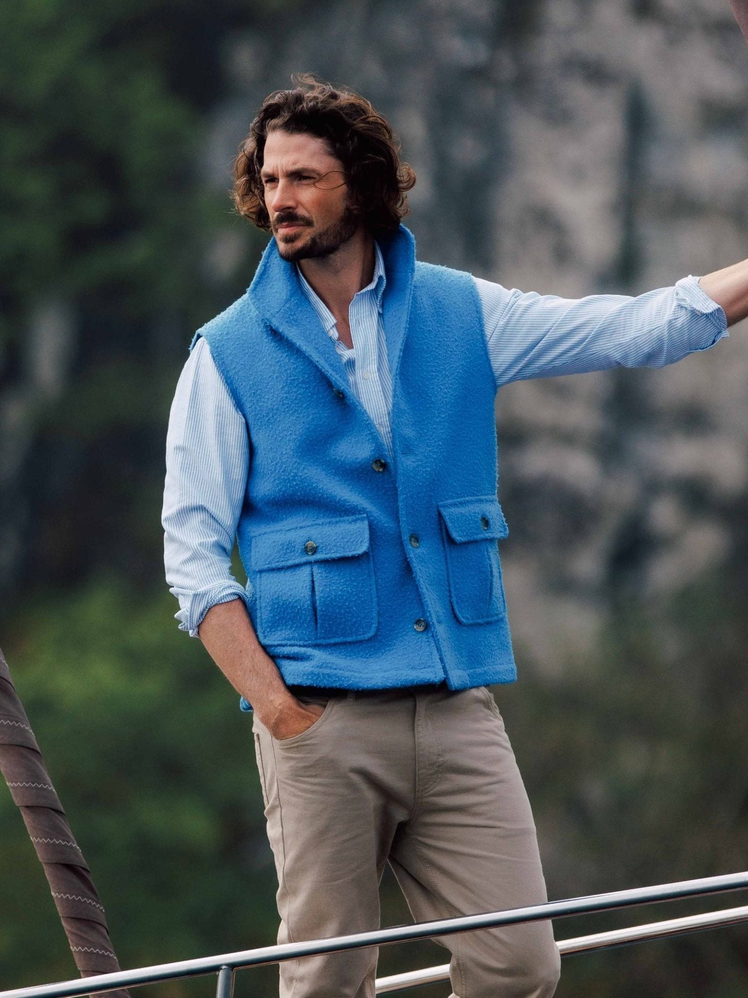 A man with wavy brown hair and a beard stands outdoors on a boat, wearing a light blue shirt, bright blue fleece vest with pockets, and Neutral Cotton Jeans from Campbells of Beauly, with a blurred natural background behind him.