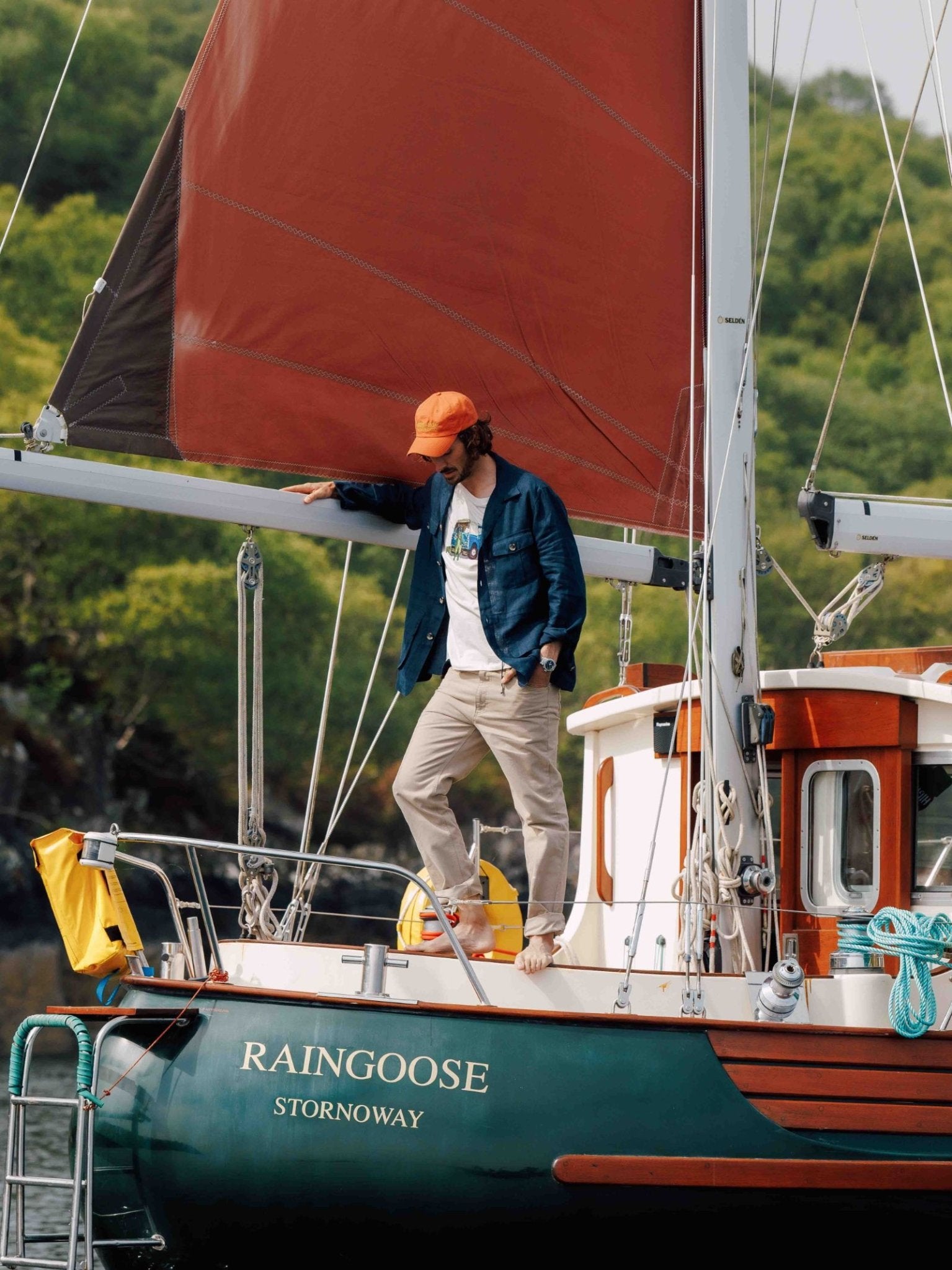 A person in a blue Campbells of Beauly Field Chore Jacket and orange cap stands barefoot on the deck of a green sailboat named RAINGOOSE STORNOWAY, surrounded by trees and greenery in the background.