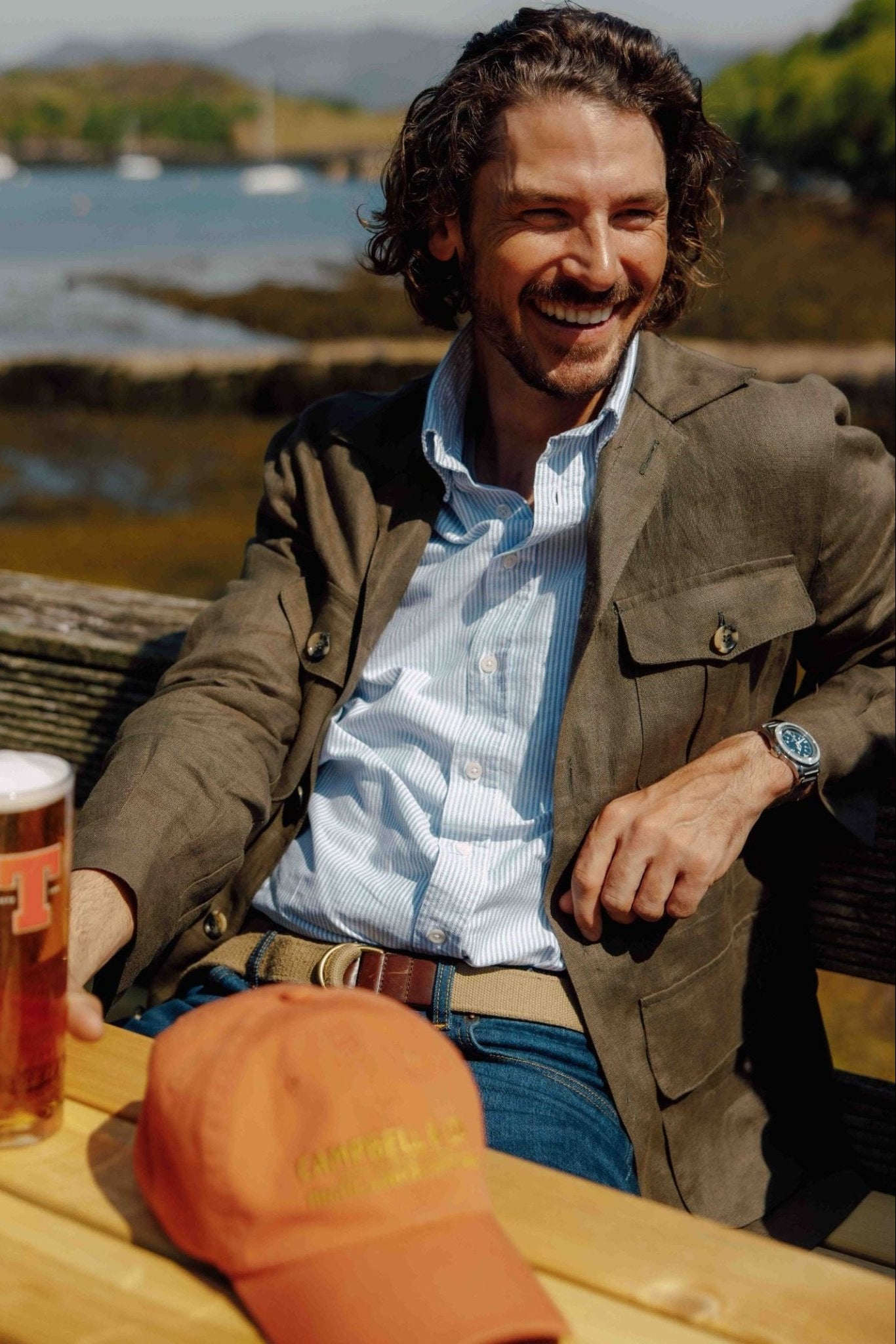 A smiling man with wavy hair sits at a wooden outdoor table, holding a pint of beer. He wears a brown jacket, blue shirt, and the Campbells of Beauly Loop Belt. An orange cap is on the table; water and hills are in the background.