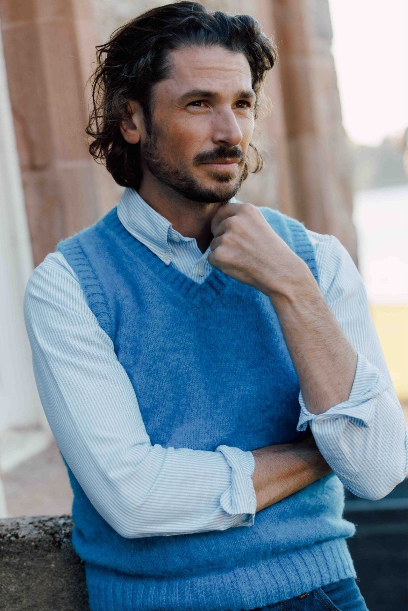 A man with wavy brown hair and a beard stands outside, thoughtfully resting his chin on his hand while wearing the Campbells of Beauly Shetland Slipover over a striped shirt.
