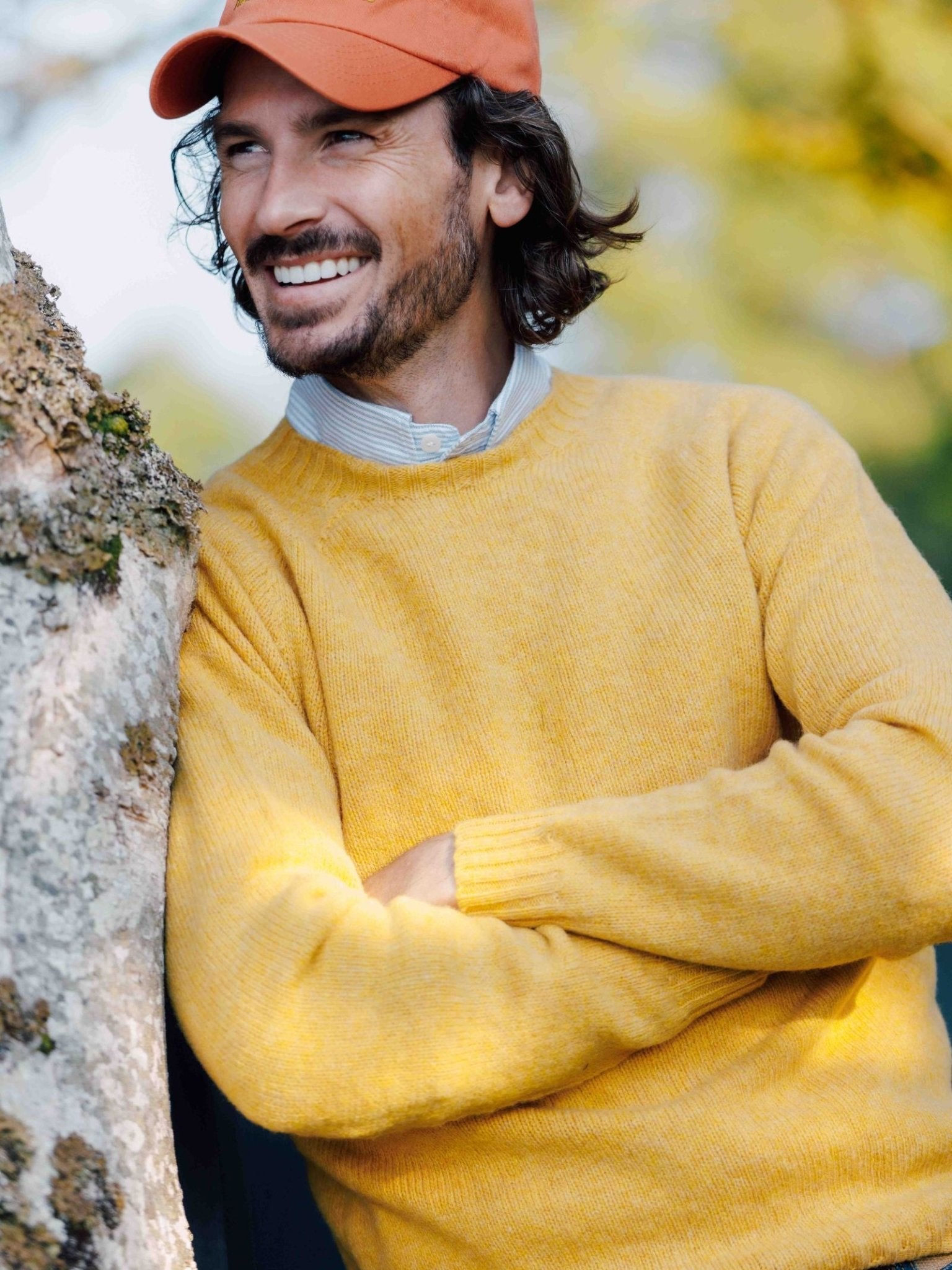 A bearded man in an orange cap, yellow sweater, and light blue shirt smiles outdoors, his style elevated by the classic Shetland Jumper from Campbells of Beauly in heritage-inspired colours.
