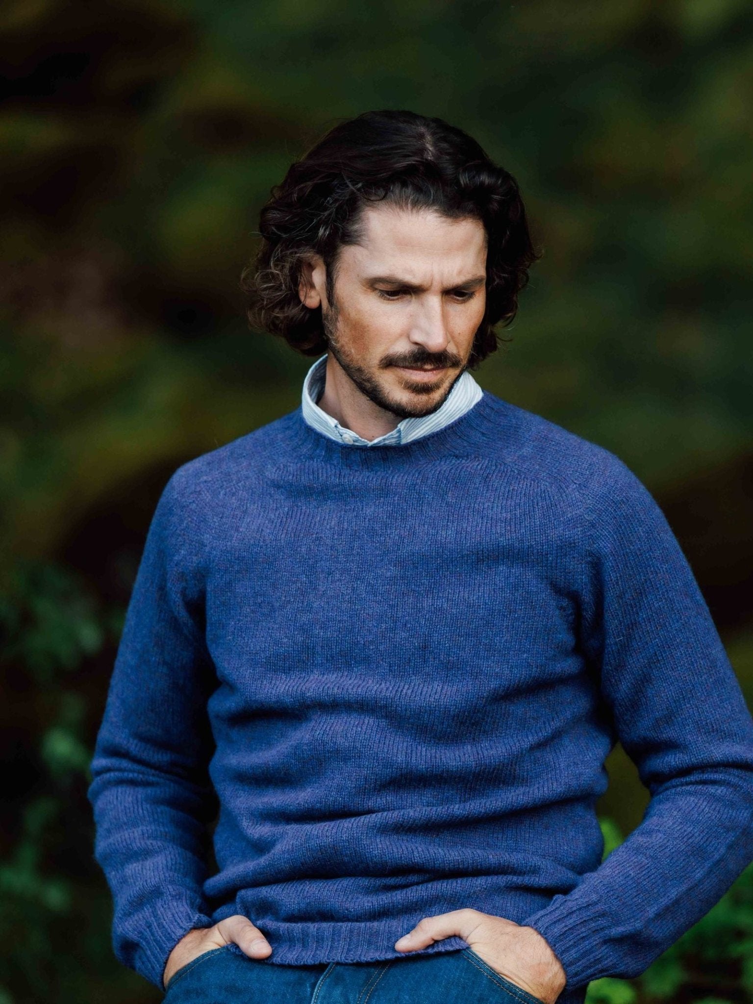 A man with wavy brown hair and a trimmed beard stands outdoors against a blurred green background, hands in pockets, wearing a blue Campbells of Beauly Shetland Jumper over a collared shirt, crafted from soft lambswool yarns.