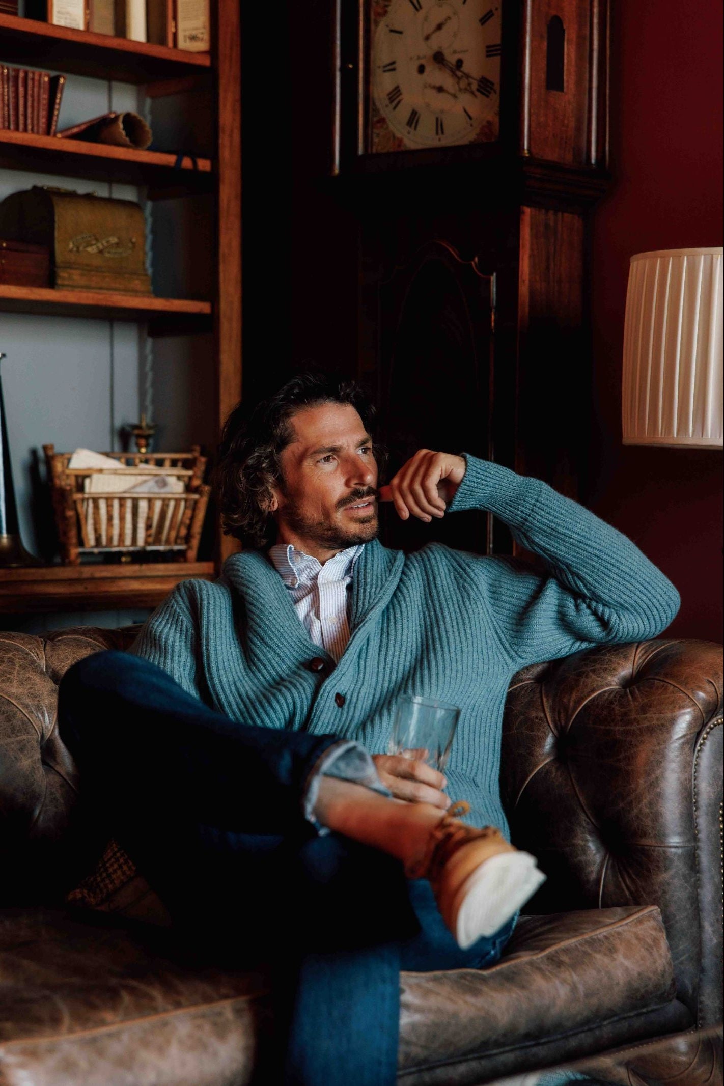 A man with wavy hair sits on a brown leather sofa wearing the Campbells of Beauly Shawl Collar Cardigan over a white shirt and jeans. He holds a glass and looks to the side, with shelves and a large clock in the background.