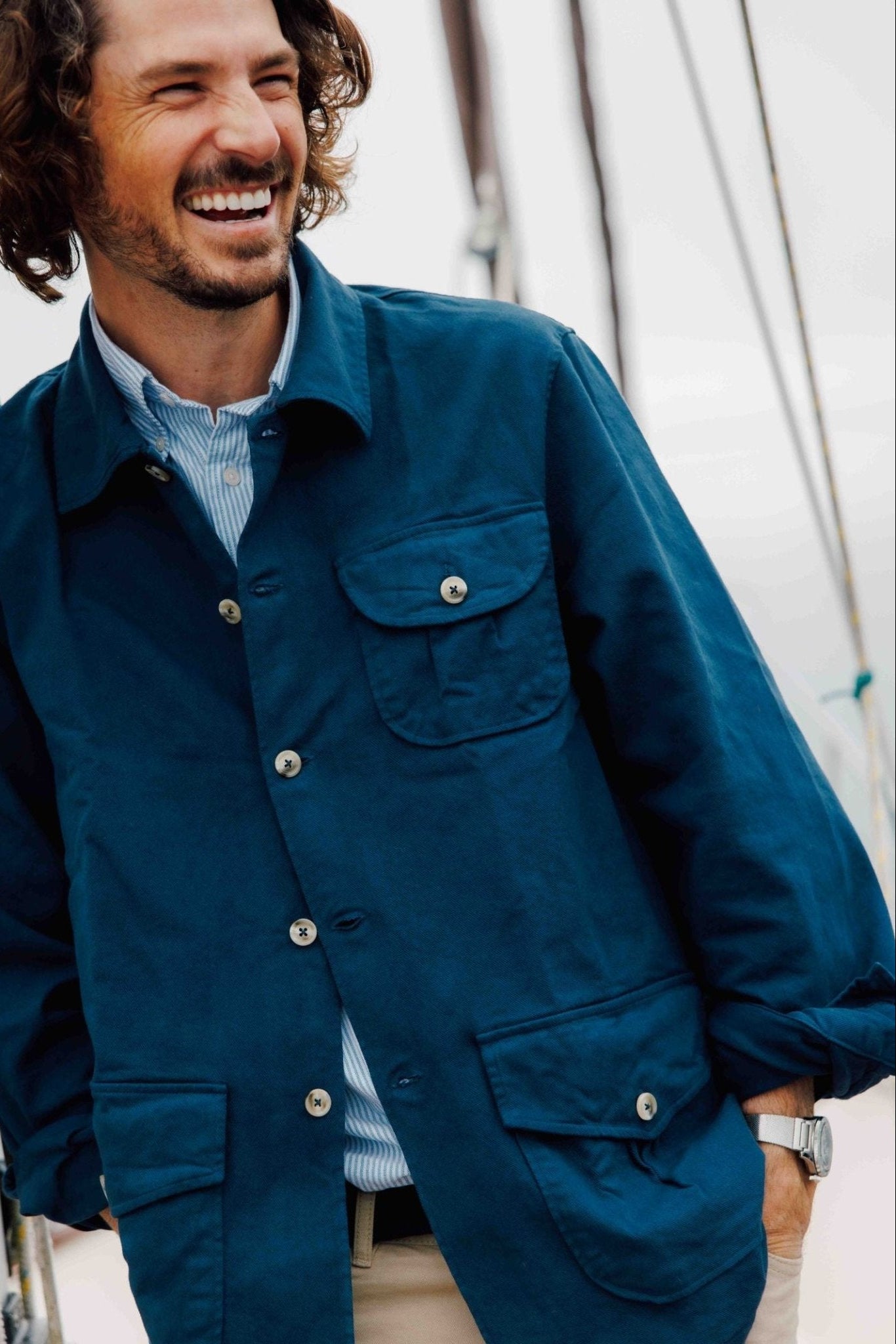 A smiling man with wavy brown hair and a beard leans on a boat railing, wearing the Campbells of Beauly Field Chore Jacket over a collared shirt and beige pants, in an outdoor setting with a blurred background.