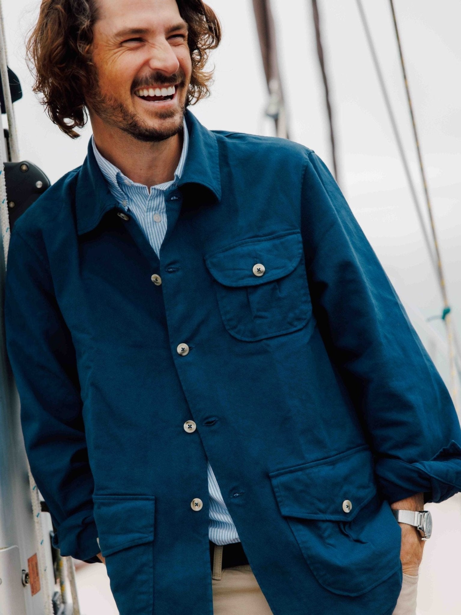 A smiling man with wavy brown hair and a beard leans on a boat railing, wearing the Campbells of Beauly Field Chore Jacket over a collared shirt and beige pants, in an outdoor setting with a blurred background.