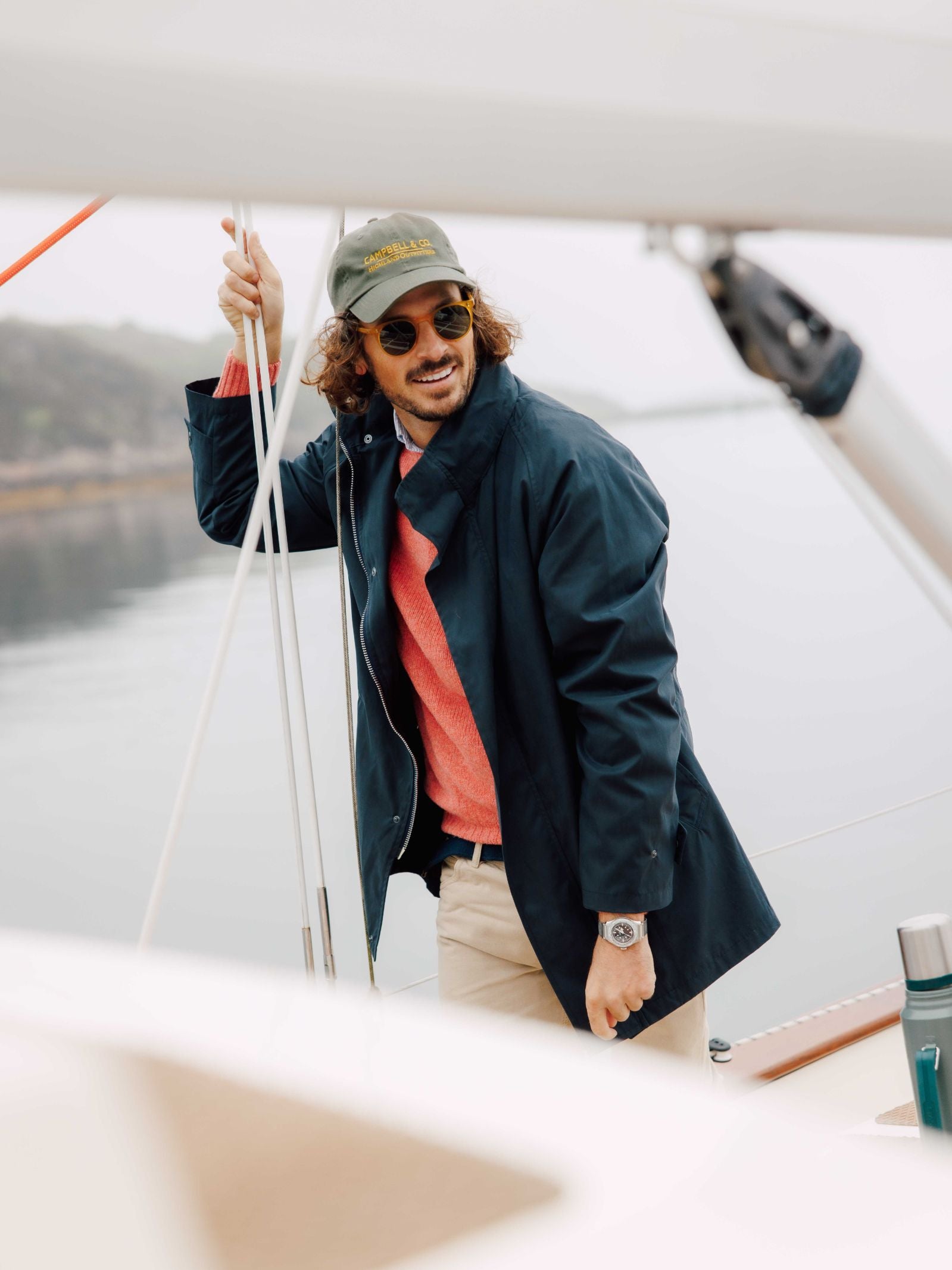 A man wearing the Campbells of Beauly Mont Coat in navy, sunglasses, a green cap, coral sweater, and beige pants stands smiling on a sailboat holding a rope with blurred calm water and trees in the background.