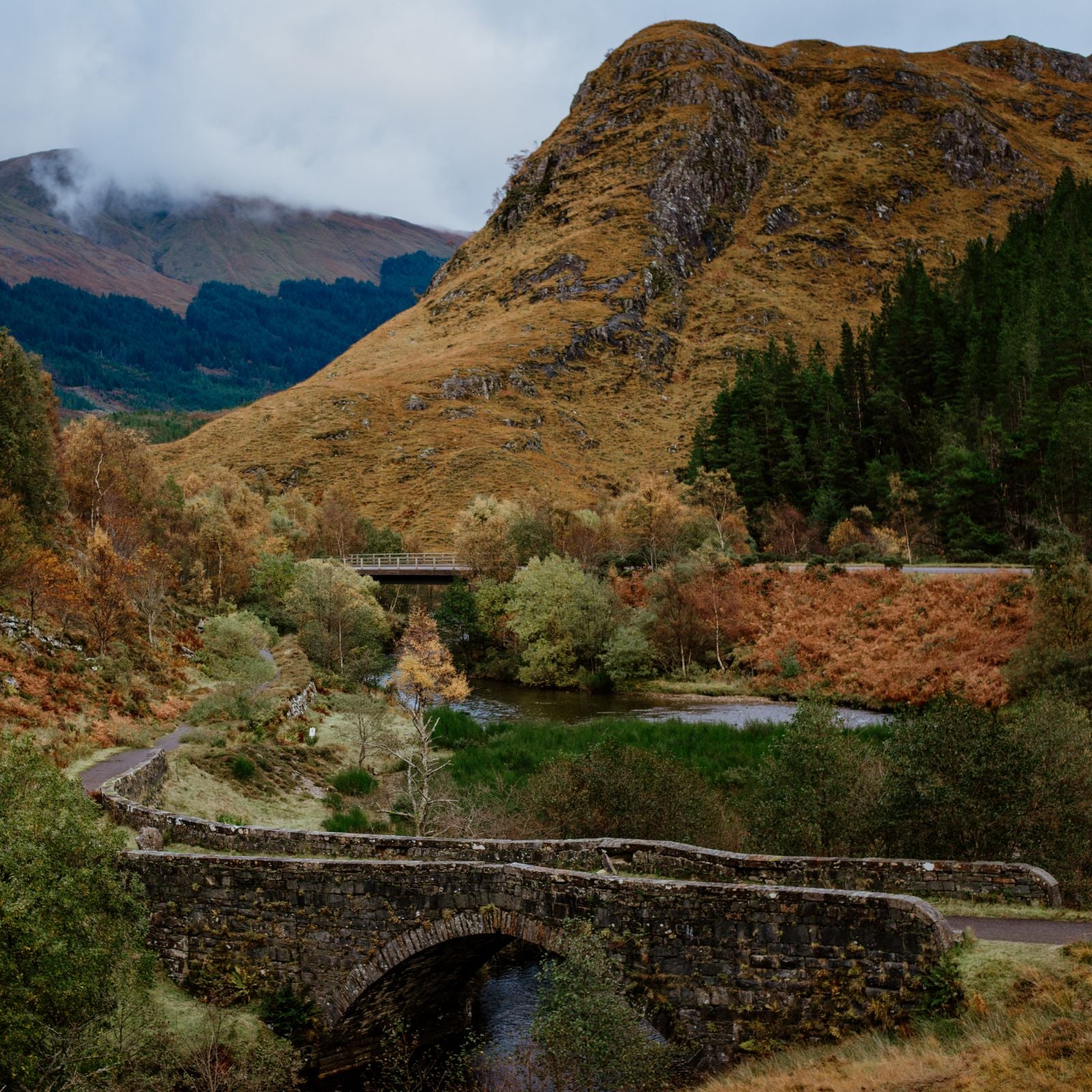 A stone bridge crosses a stream in a scenic, hilly landscape with autumnal foliage, green trees, and a tall, rocky hill in the background under a cloudy sky.