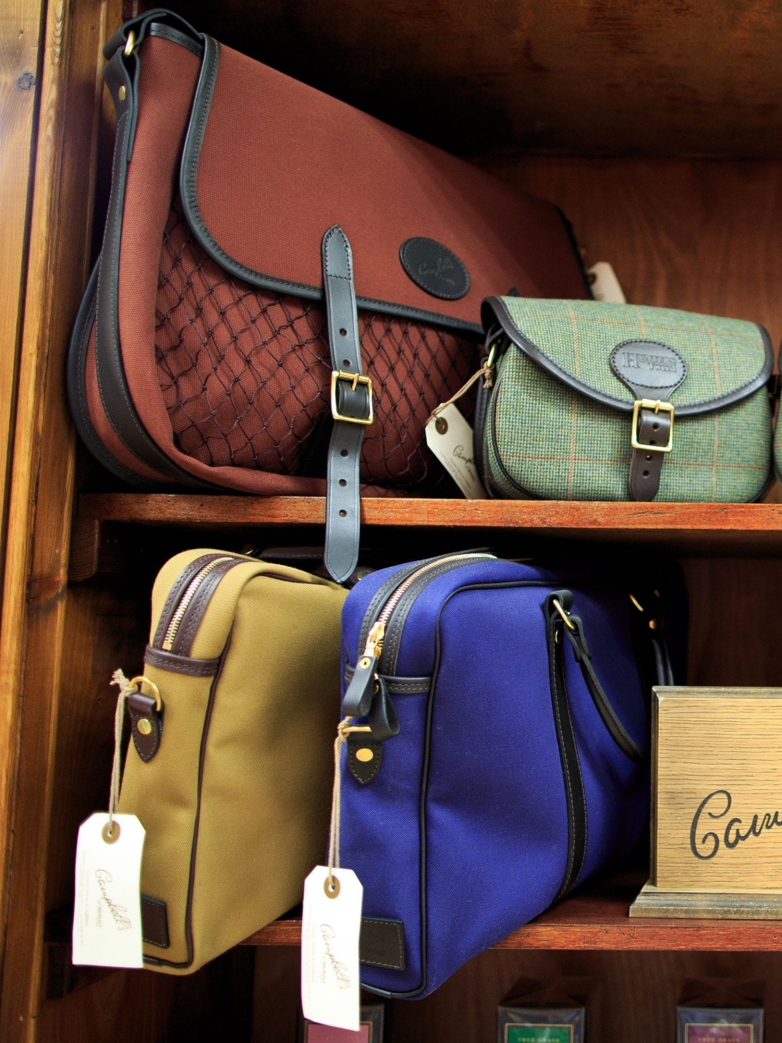 Four stylish Campbell's of Beauly Laptop Bags are displayed on wooden shelves, featuring buckles, tags, and leather accents in brown, green, tan, and blue—including canvas and business styles.