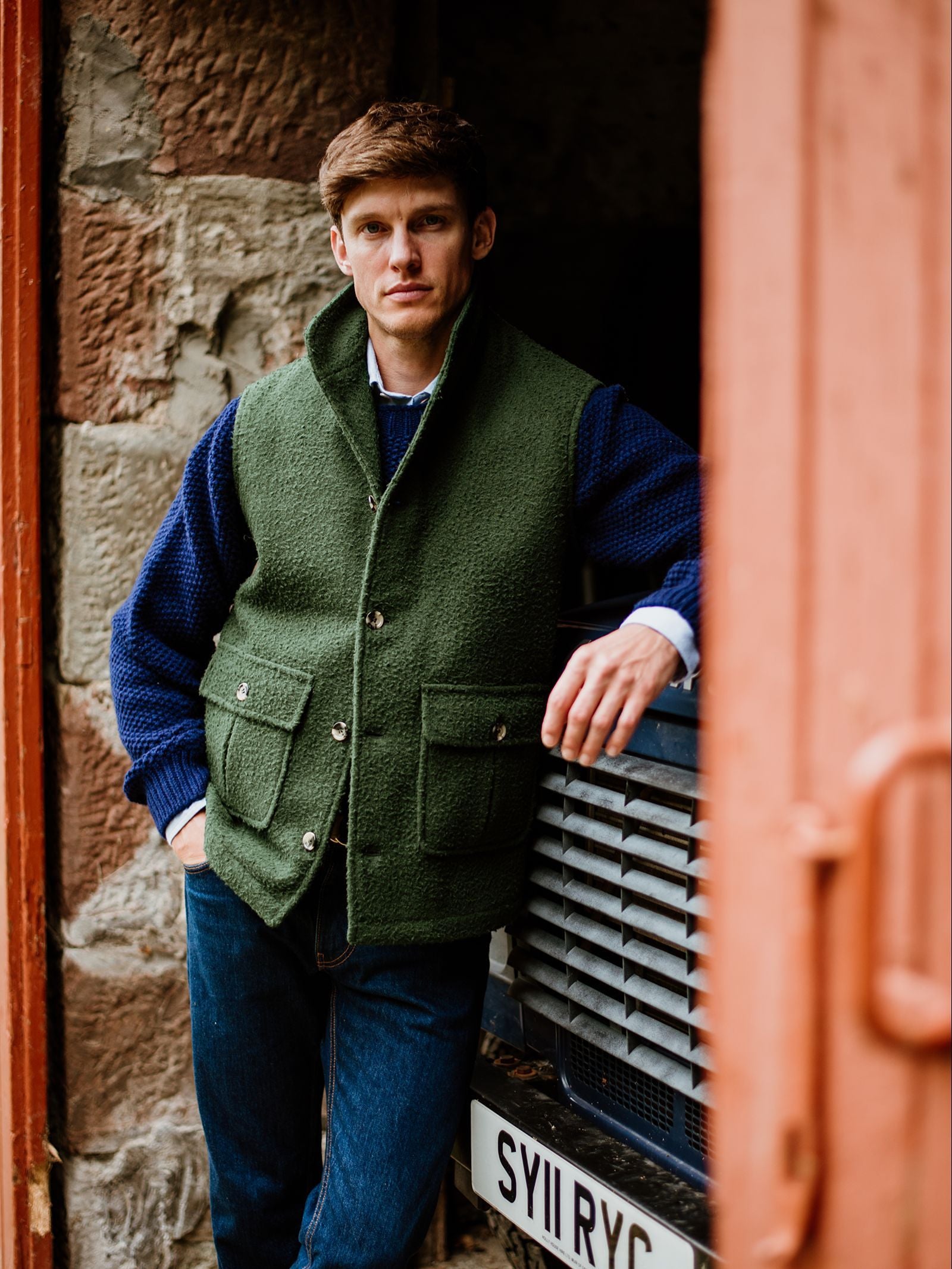 A man wearing a green Campbell’s of Beauly Casentino Fleece Gilet and a blue sweater leans against a dark vehicle with a visible license plate, standing in a rustic doorway adorned with stone and wood details.