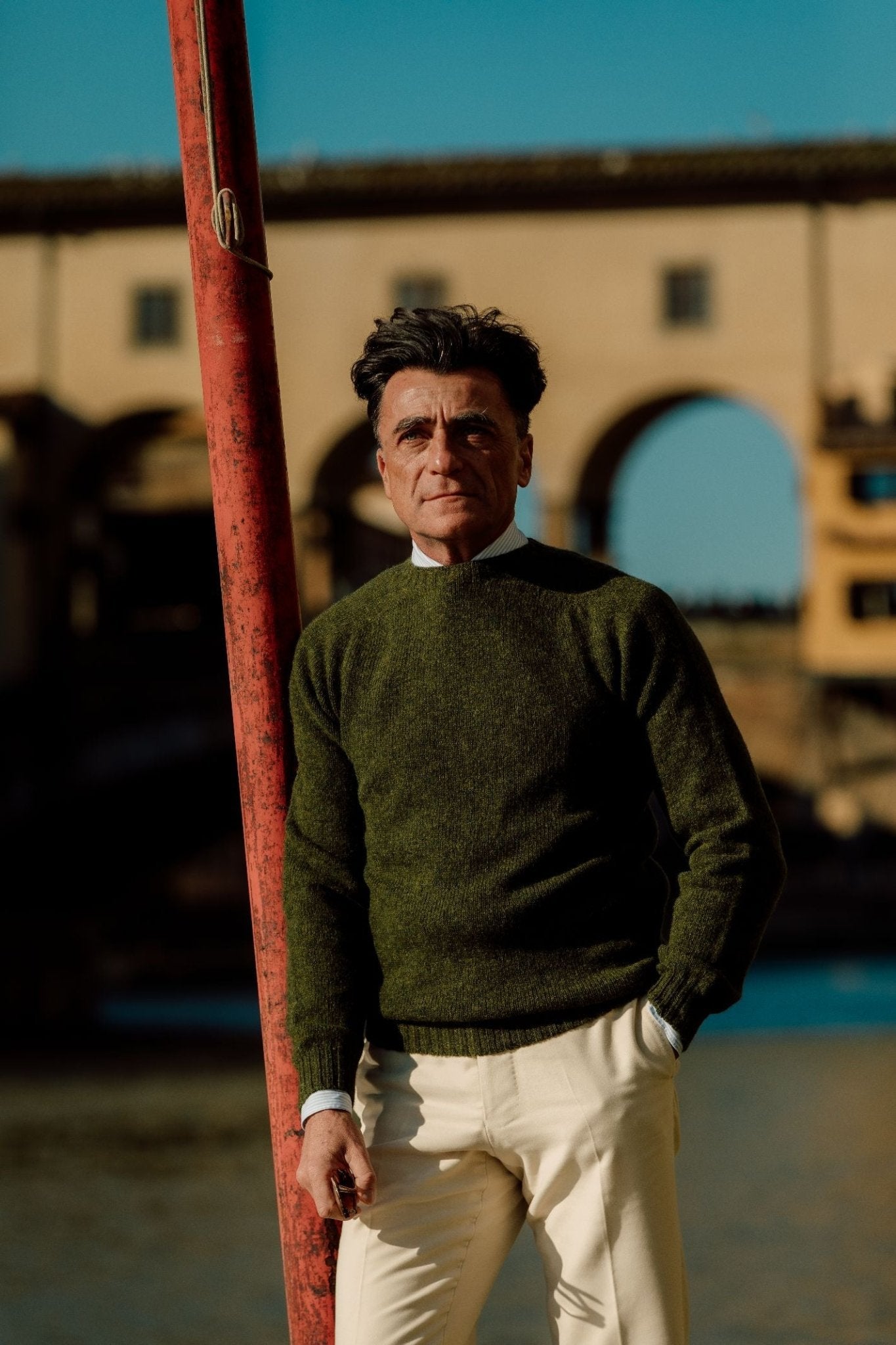 A man stands outdoors by a red pole, wearing a green Campbells of Beauly Shetland Jumper with light pants; an arched stone bridge and blue sky are in the background.