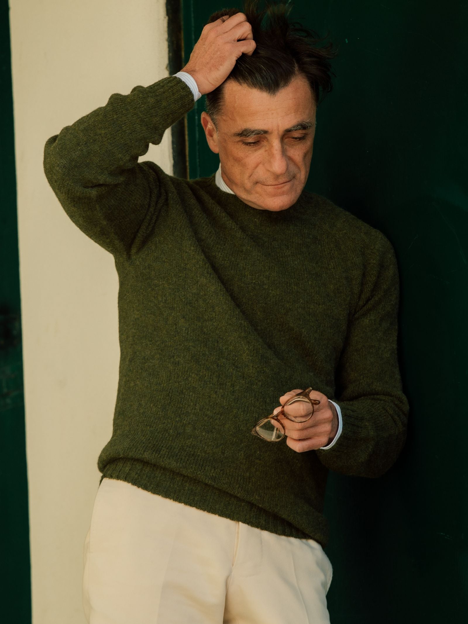 A man in a Campbell's of Beauly Shetland Jumper and cream pants stands against a dark green door, holding eyeglasses in one hand, brushing his hair with the other, and gazing down thoughtfully.
