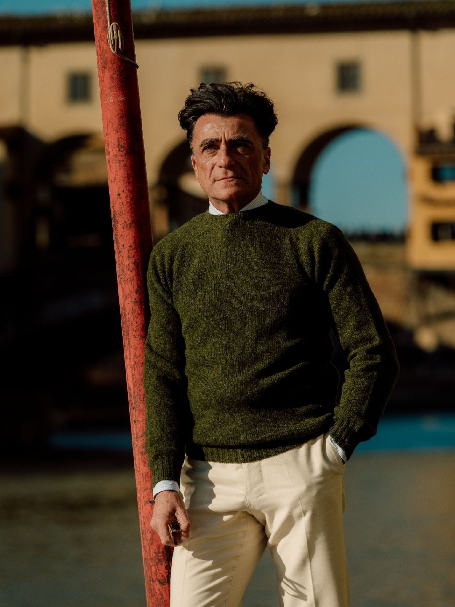 A man with dark hair, wearing a Campbell's of Beauly Shetland Jumper in olive green and cream pants, stands by a red pole near a river, with an old stone bridge and buildings in the background under a clear blue sky.
