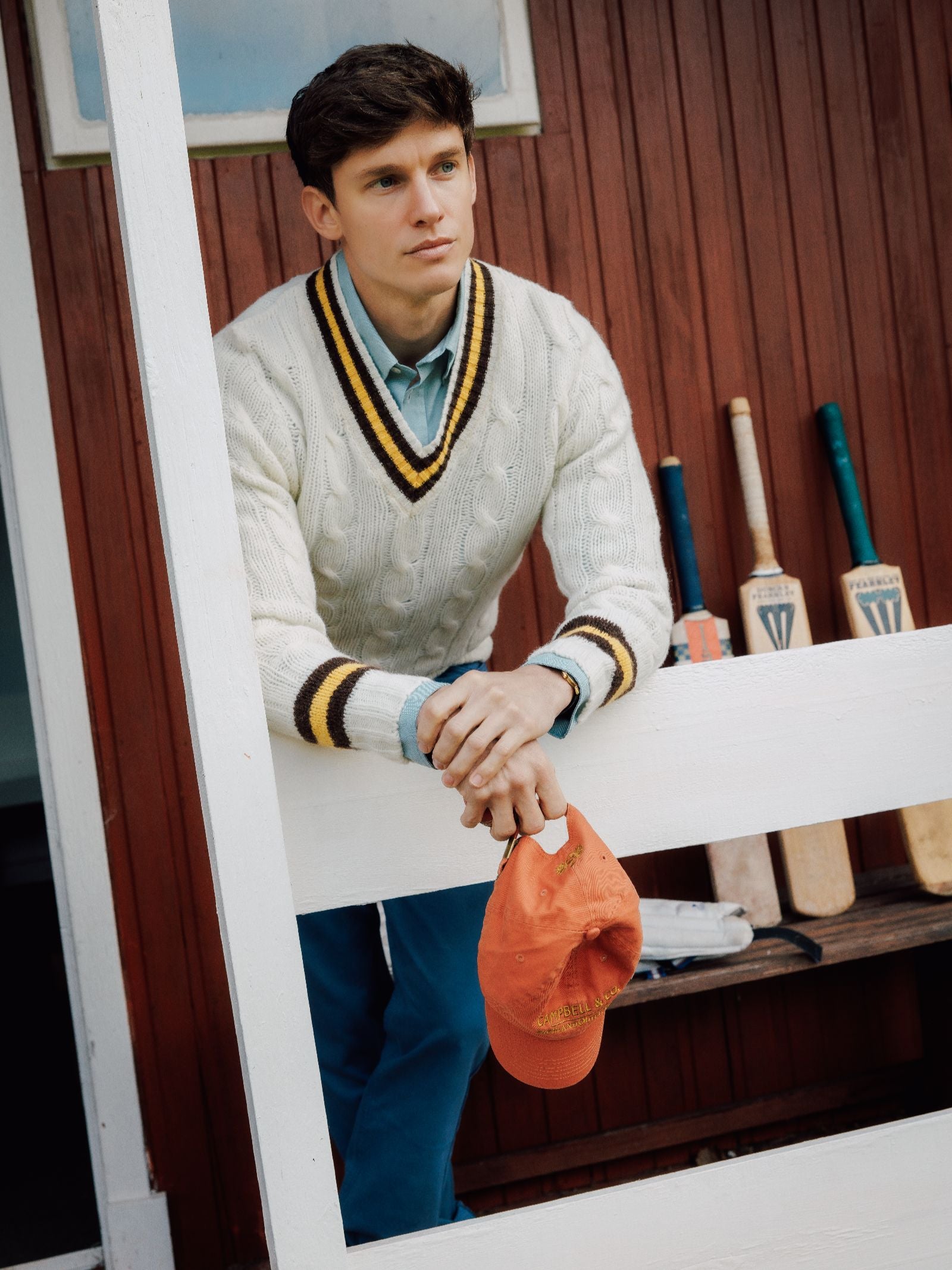 A young man in the Campbell's of Beauly Shetland Cricket Sweater and blue pants leans on a white fence, holding an orange cap. Cricket bats rest against a red wall, evoking timeless cricket style.