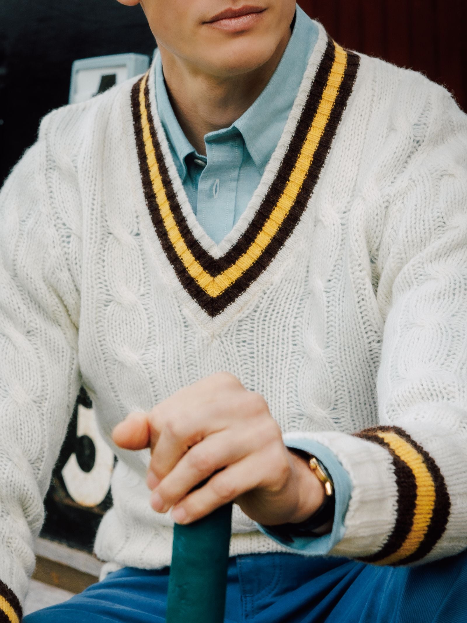 Wearing the Campbell's of Beauly Shetland Cricket Sweater with yellow and brown trim over a light blue shirt, a person grips a green handle. The cropped photo shows only the lower face and torso, highlighting classic cricket style.