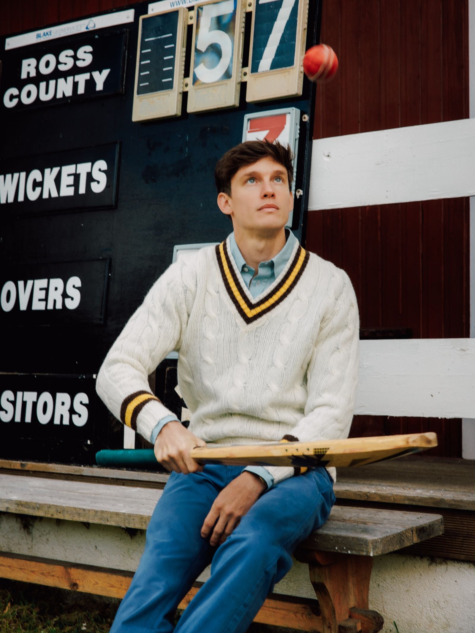 A young man in a cream Campbell's of Beauly Shetland Cricket Sweater and blue pants sits on a bench, tossing a cricket ball and holding a bat. Behind him, the scoreboard shows “Ross County” with a score of 157.