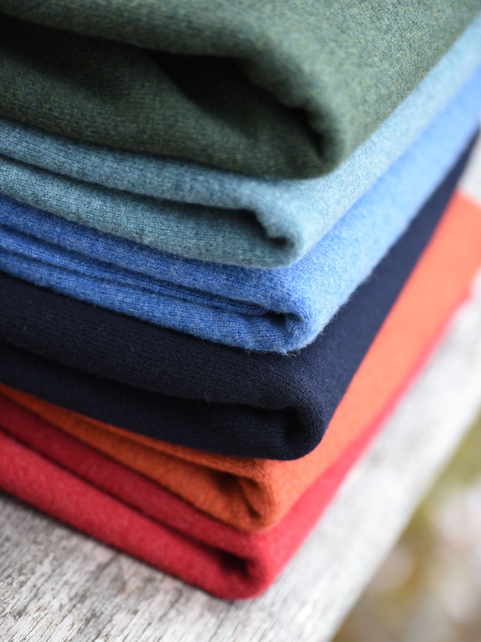 A neatly stacked pile of folded Campbells of Beauly Lambswool Crew Neck Jumpers in green, light blue, navy, orange, and red rests on a weathered wooden surface, highlighting their soft texture and vibrant colors.