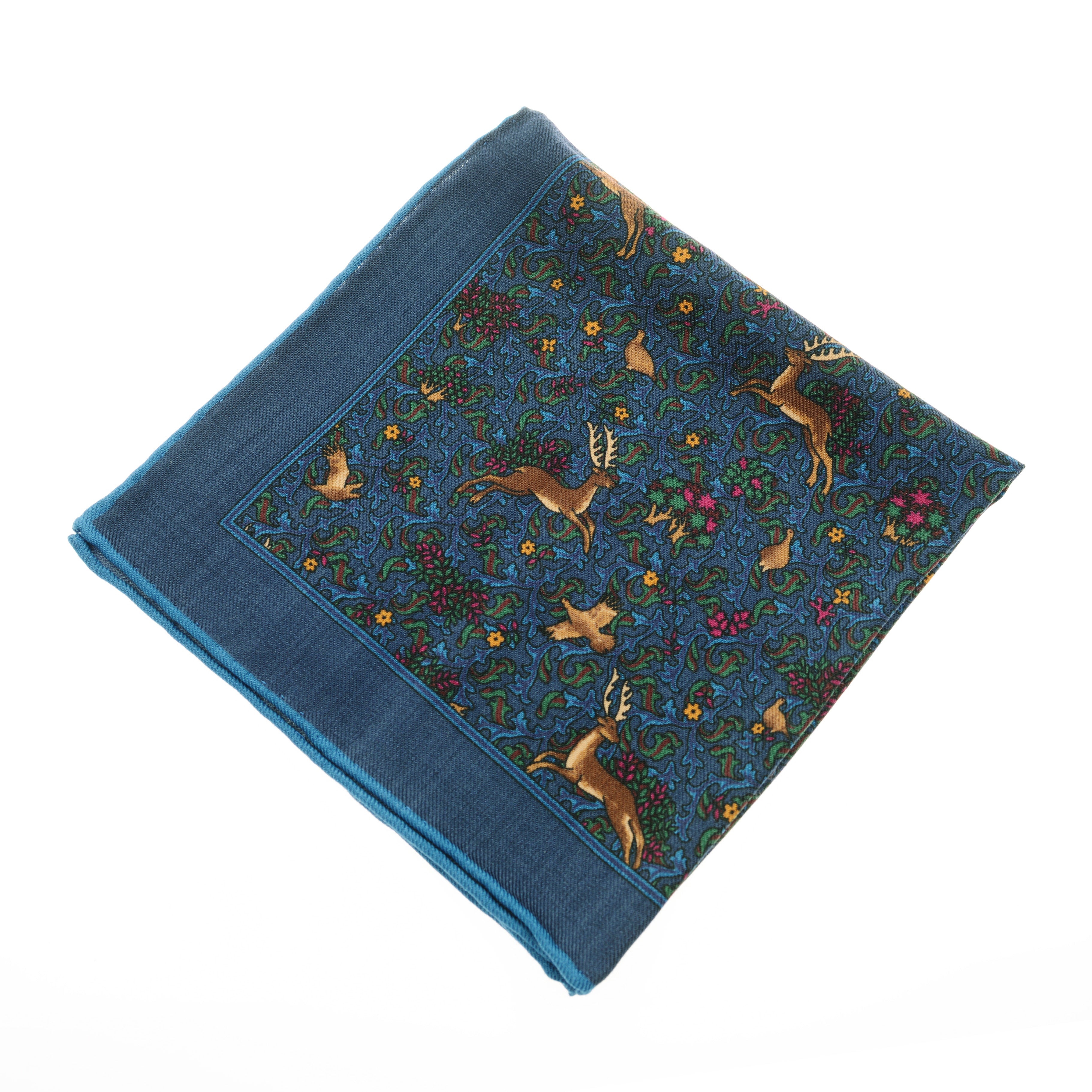 The Wool Hank by Campbells of Beauly is a blue wool handkerchief with hand-rolled edges, displaying deer and rabbits among green vines, pink flowers, and yellow blooms on a rich blue background with a decorative border.