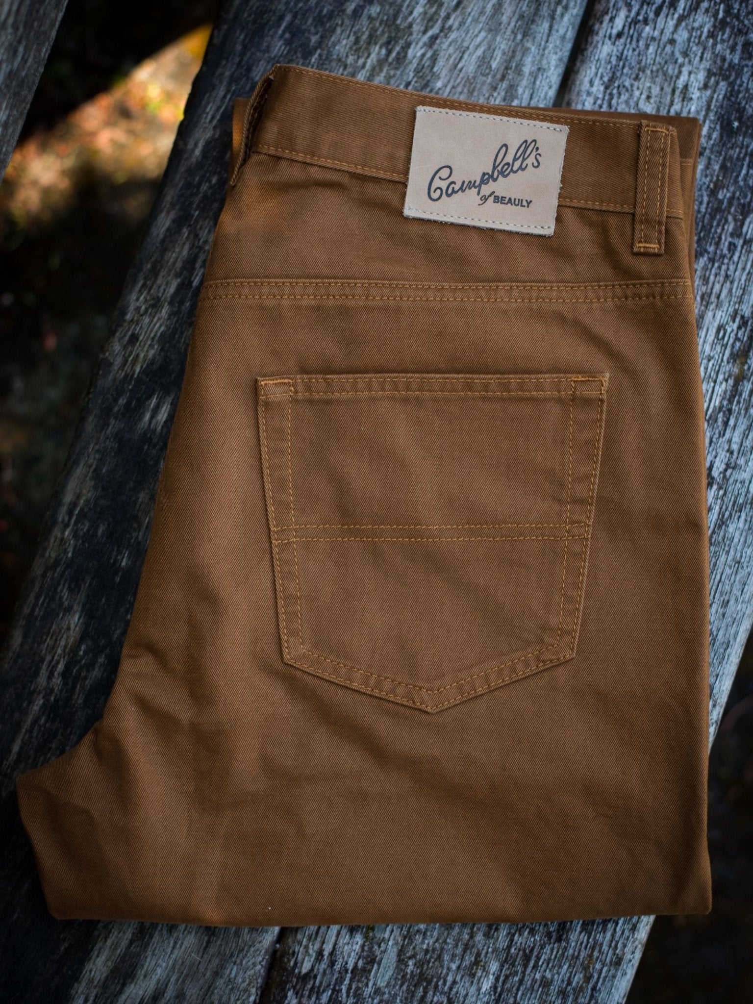 A pair of folded brown Colour Cotton Jeans by Campbell's of Beauly, featuring a straight leg and back pocket, is displayed on a weathered wooden surface.