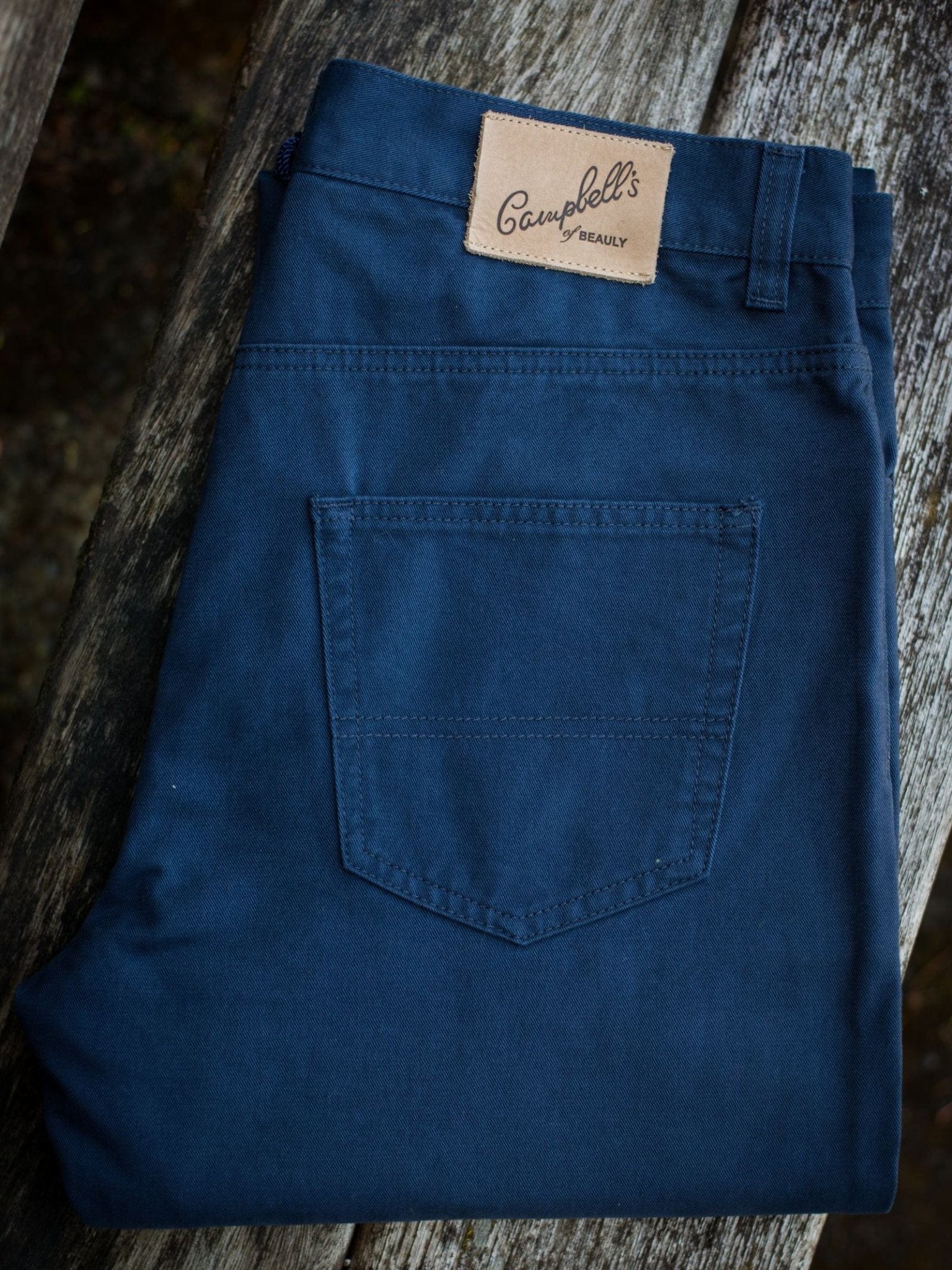 A pair of folded dark blue Colour Cotton Jeans by Campbell's of Beauly, featuring a five-pocket design and a leather patch above the back pocket, is displayed on a weathered wooden surface.
