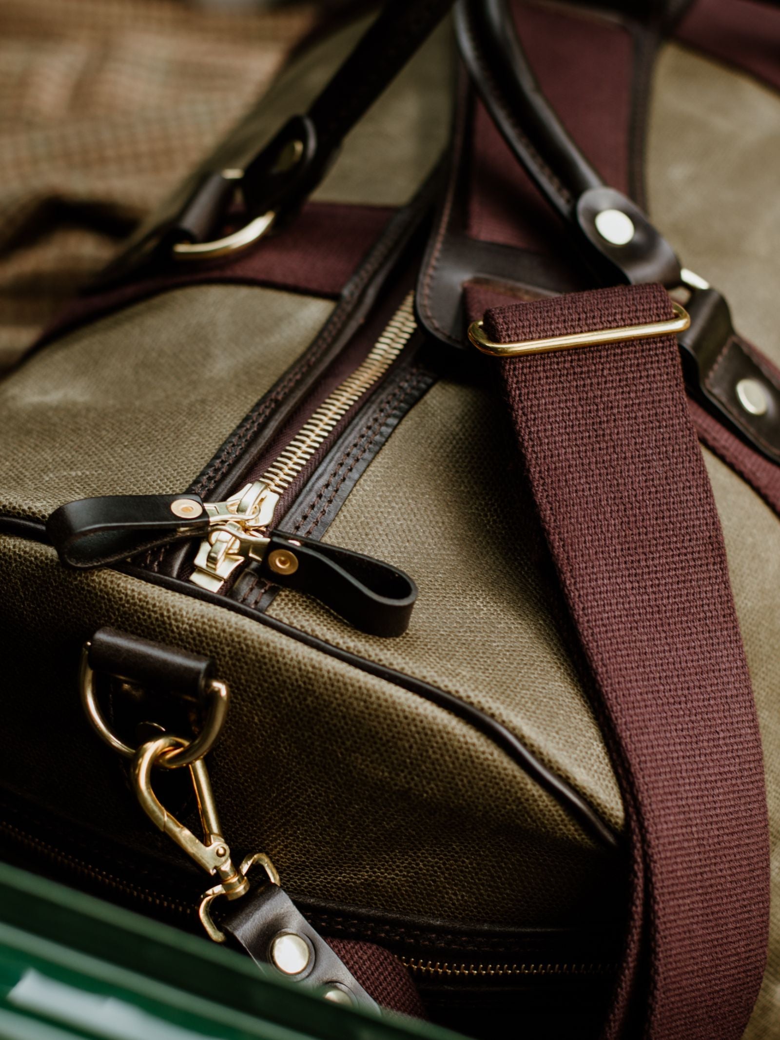 Close-up of the Campbell's of Beauly Wax Holdall: a vintage-inspired khaki canvas bag with brown leather accents, gold zipper, metal clasp, and wide maroon shoulder strap—resting on checkered fabric, it’s your ideal travel companion.