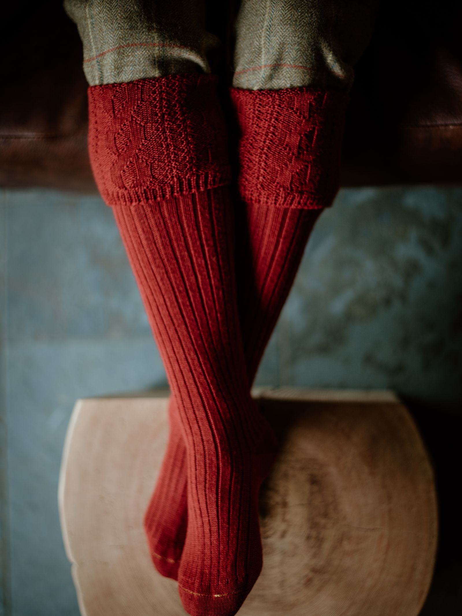 A person sits with ankles crossed, wearing Campbells of Beauly Merino Glenmore Shooting Socks and gray-green pants. Their feet rest on a round wooden stool placed on blue-gray tile, bathed in gentle natural light.