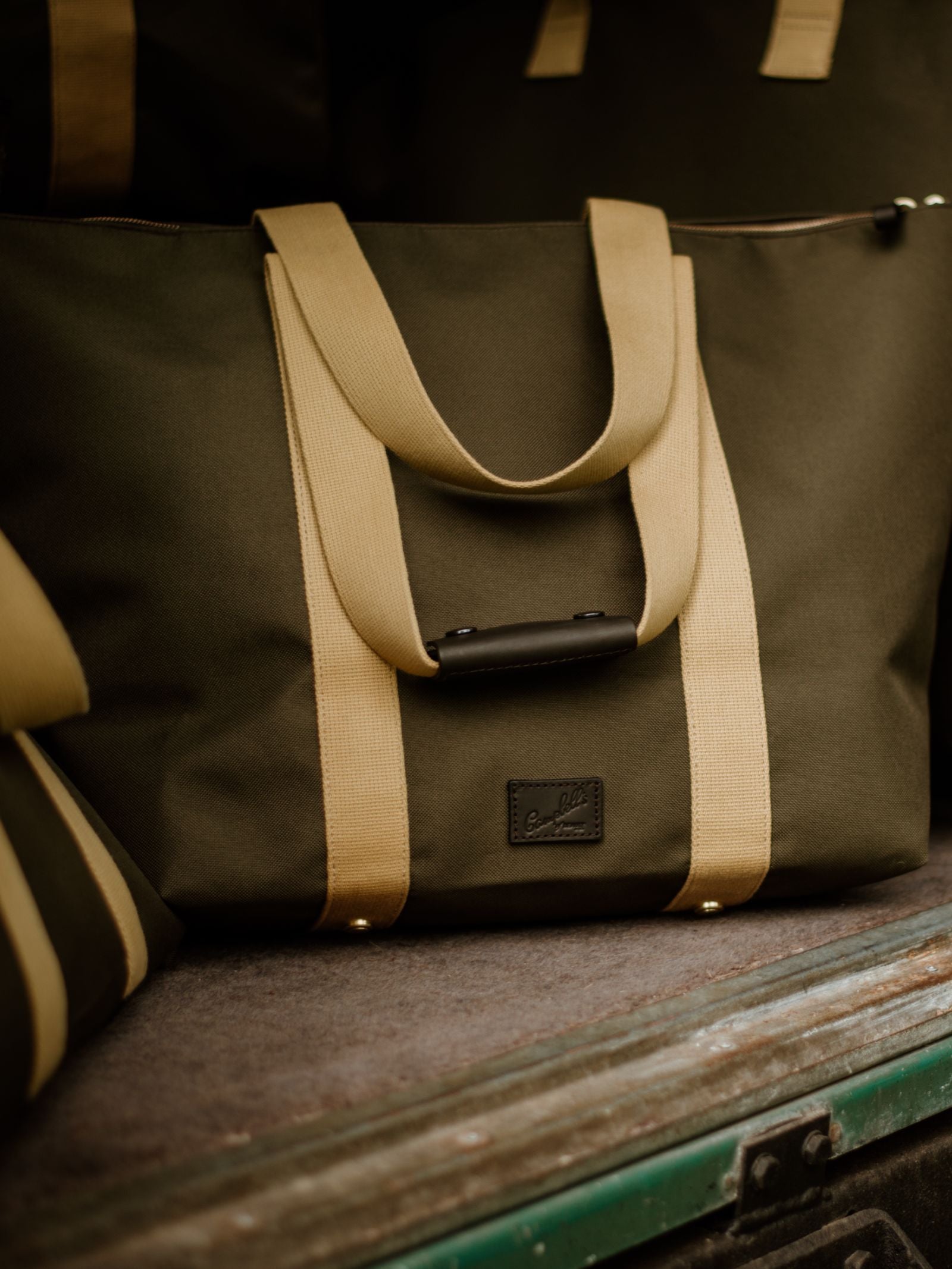 A dark green Campbell's of Beauly Utility Medium Tote with tan handles sits on a surface. Made from heavy-duty canvas, it features a black rectangular patch on the front; part of another bag is visible in the background.