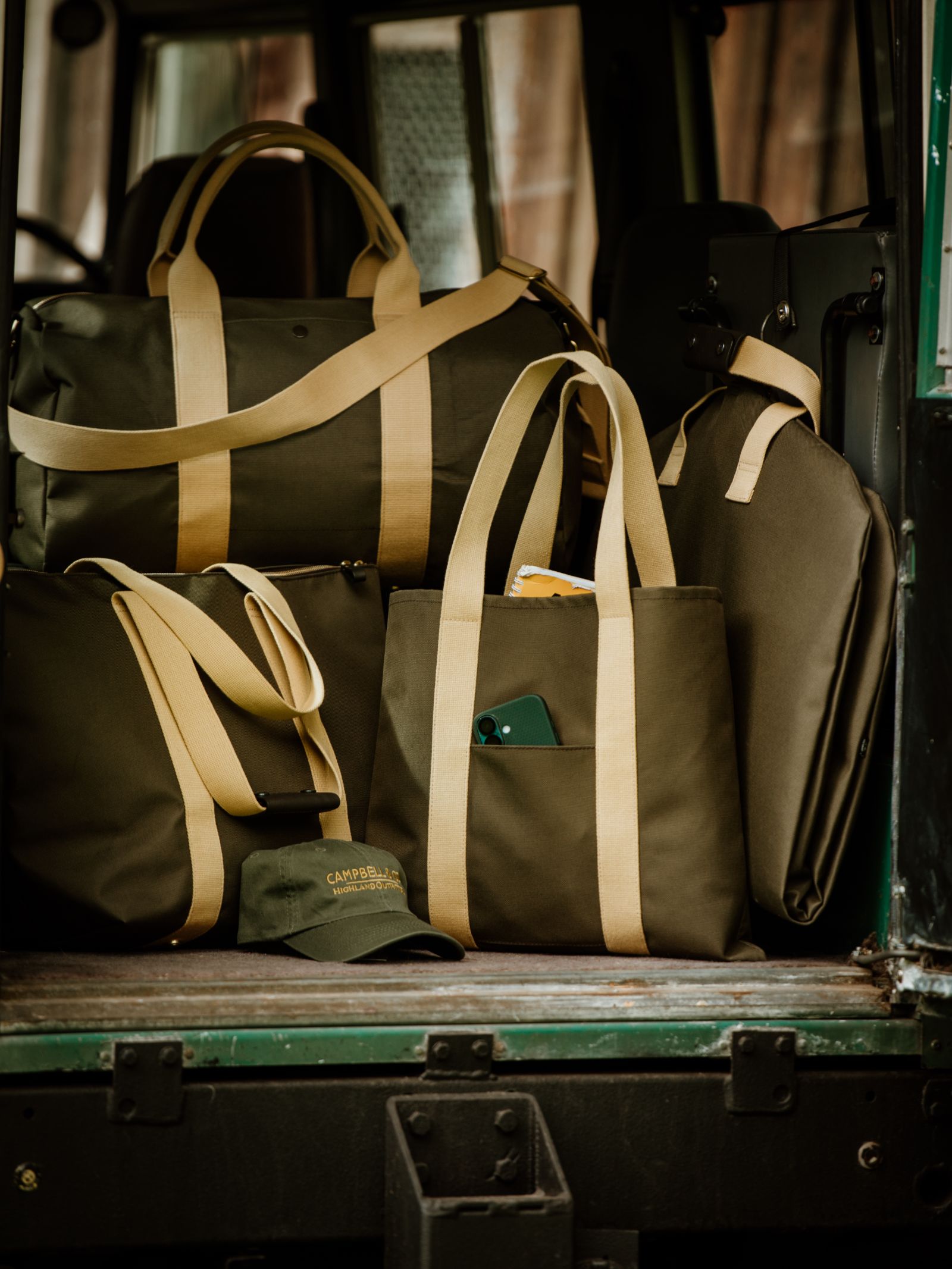 A Campbell’s of Beauly Utility Packaway Tote and a green everyday utility bag with tan straps sit on a vehicle, one holding a green smartphone in its pocket, while the background stays blurred.