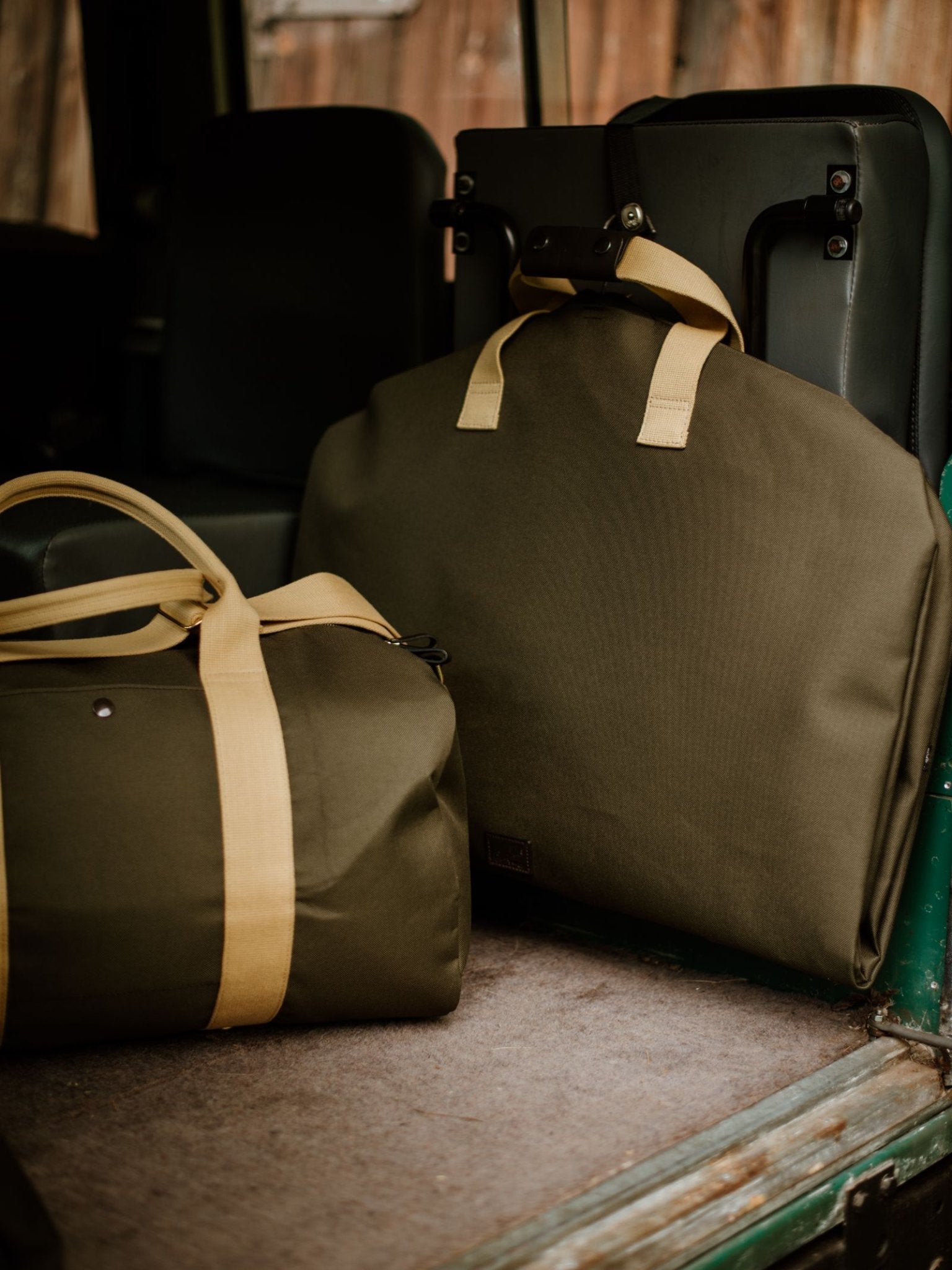 Two Campbell's of Beauly Utility Suit Bags in olive green canvas with tan straps rest on the back seat of a vehicle, featuring dark upholstery and a wooden panel in the background.