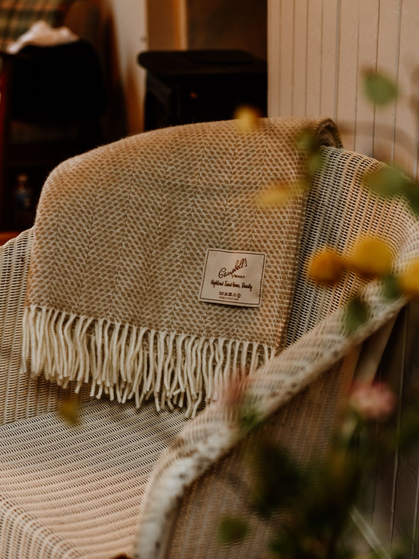 A Campbell's of Beauly Stipled Twill Blanket in beige with white fringe is draped over a wicker chair, label visible. Soft lighting and blurred yellow flowers evoke a cozy, inviting mood.