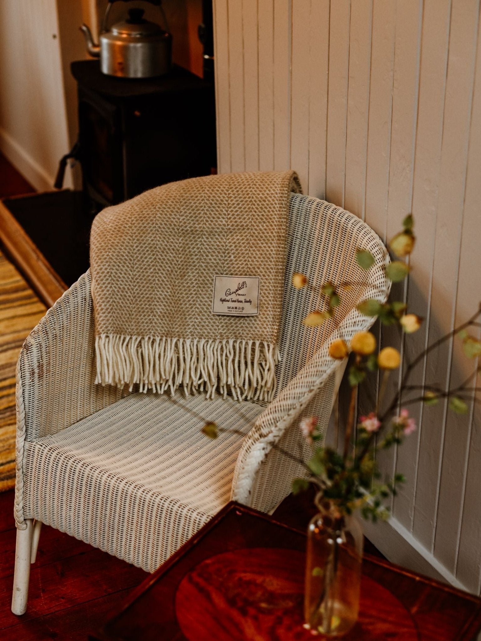 A white wicker chair with a Campbell's of Beauly Stipled Twill Blanket in beige draped over it sits next to a wooden table with flowers; in the background, a wood stove and kettle add to the cozy scene.