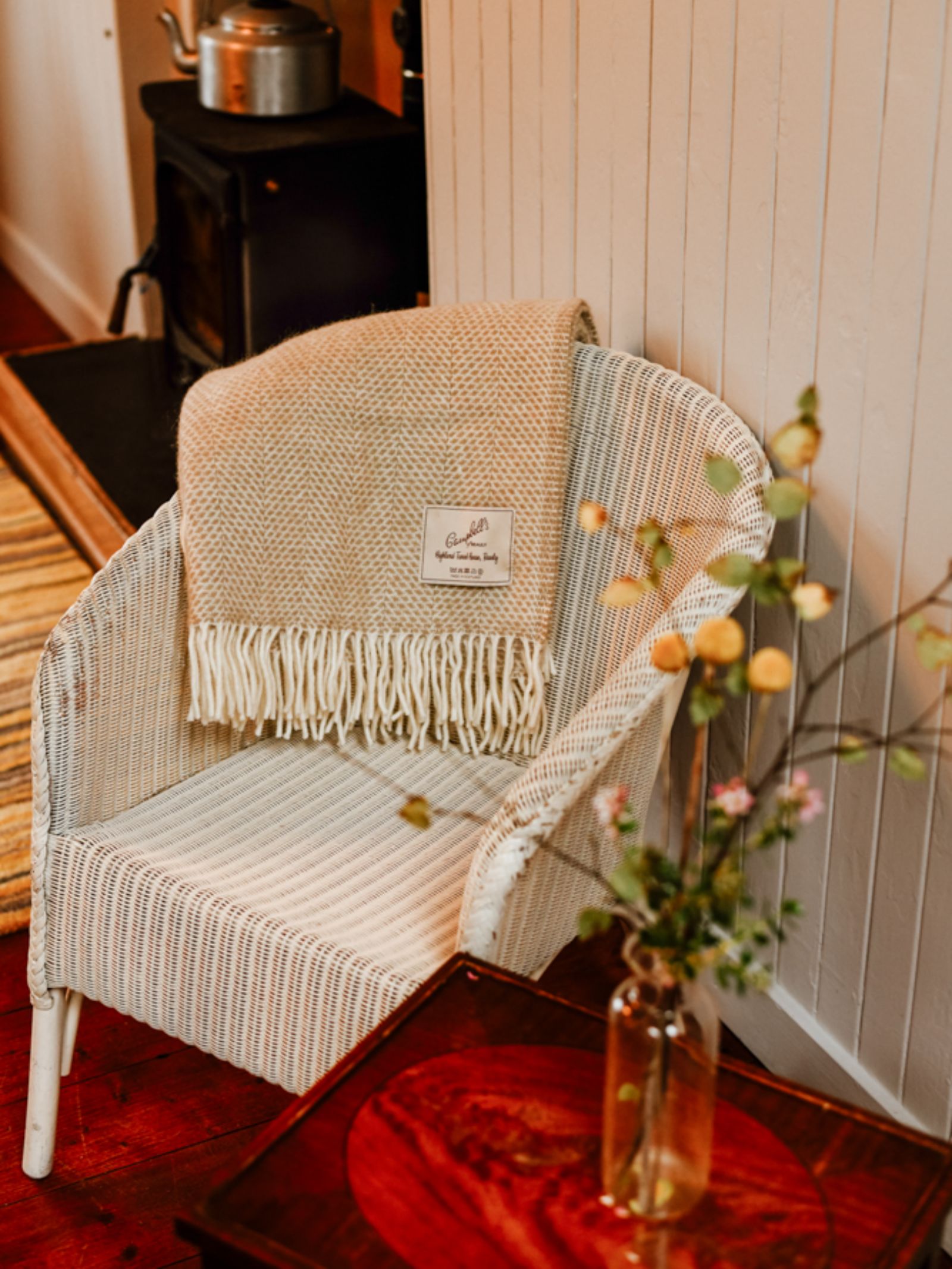 A white wicker chair with a breathable weave features the Campbell's of Beauly Stipled Twill Blanket draped over the back, beside a small wooden table with a vase of flowers. A wood stove and teapot appear in the background.