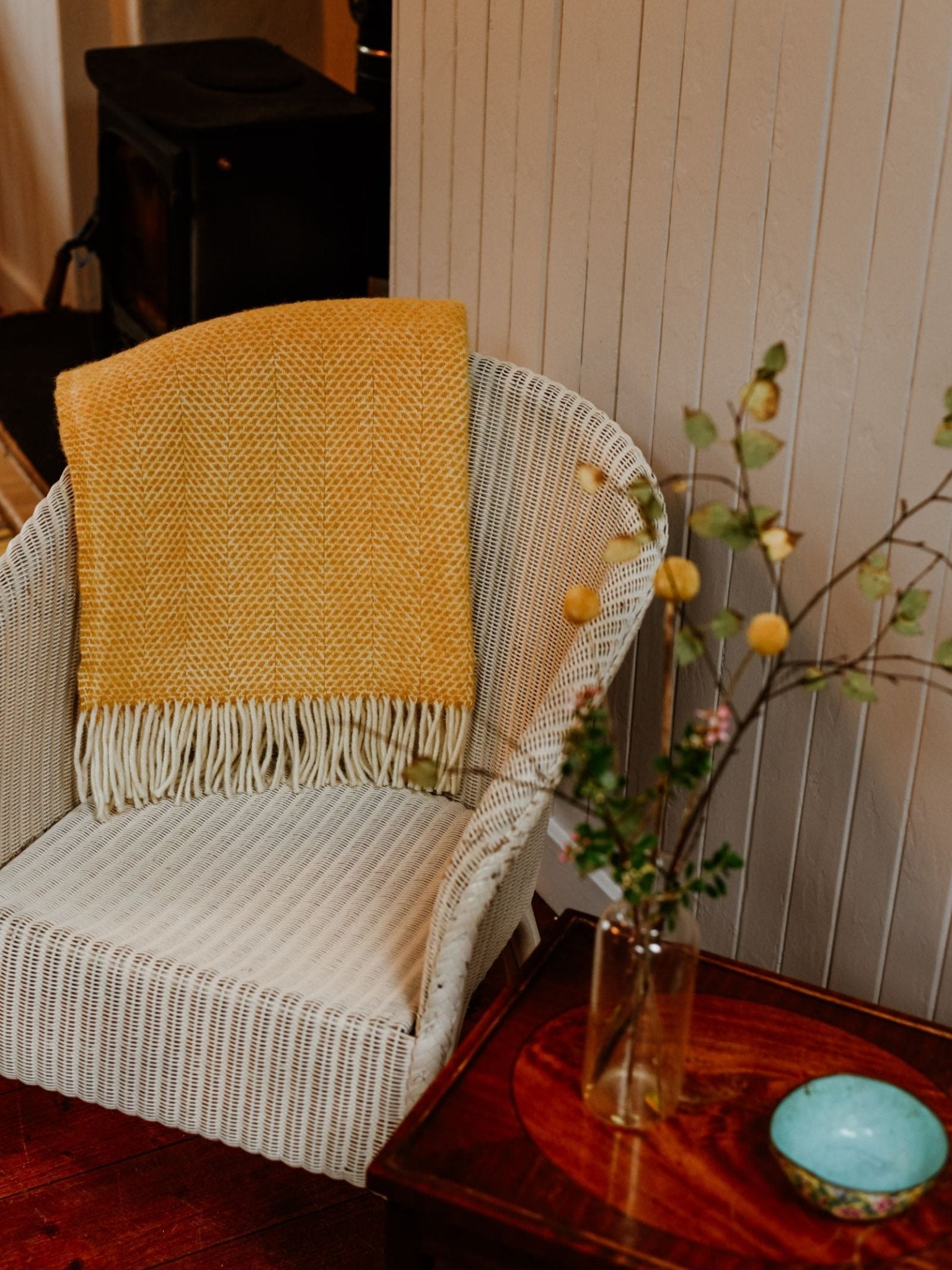 A white wicker chair with a yellow Stipled Twill Blanket from Campbell's of Beauly draped over it sits next to a wooden side table topped with a glass vase holding branches and a small turquoise bowl.