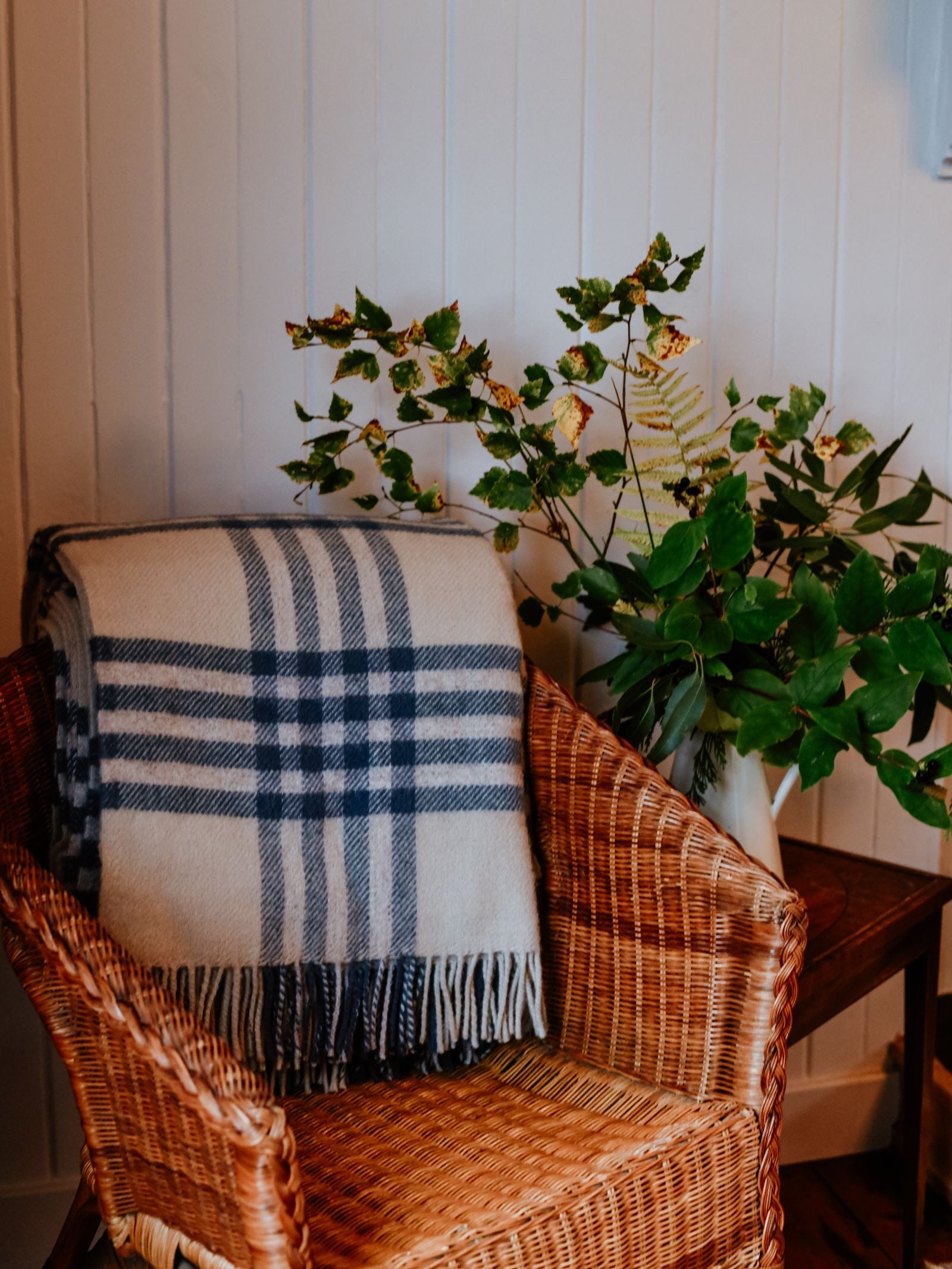 A wicker chair with a Campbell's of Beauly Tarradale Lambswool Throw draped over the back sits next to a small table holding a vase of green and yellow leafy branches, set against a white paneled wall.