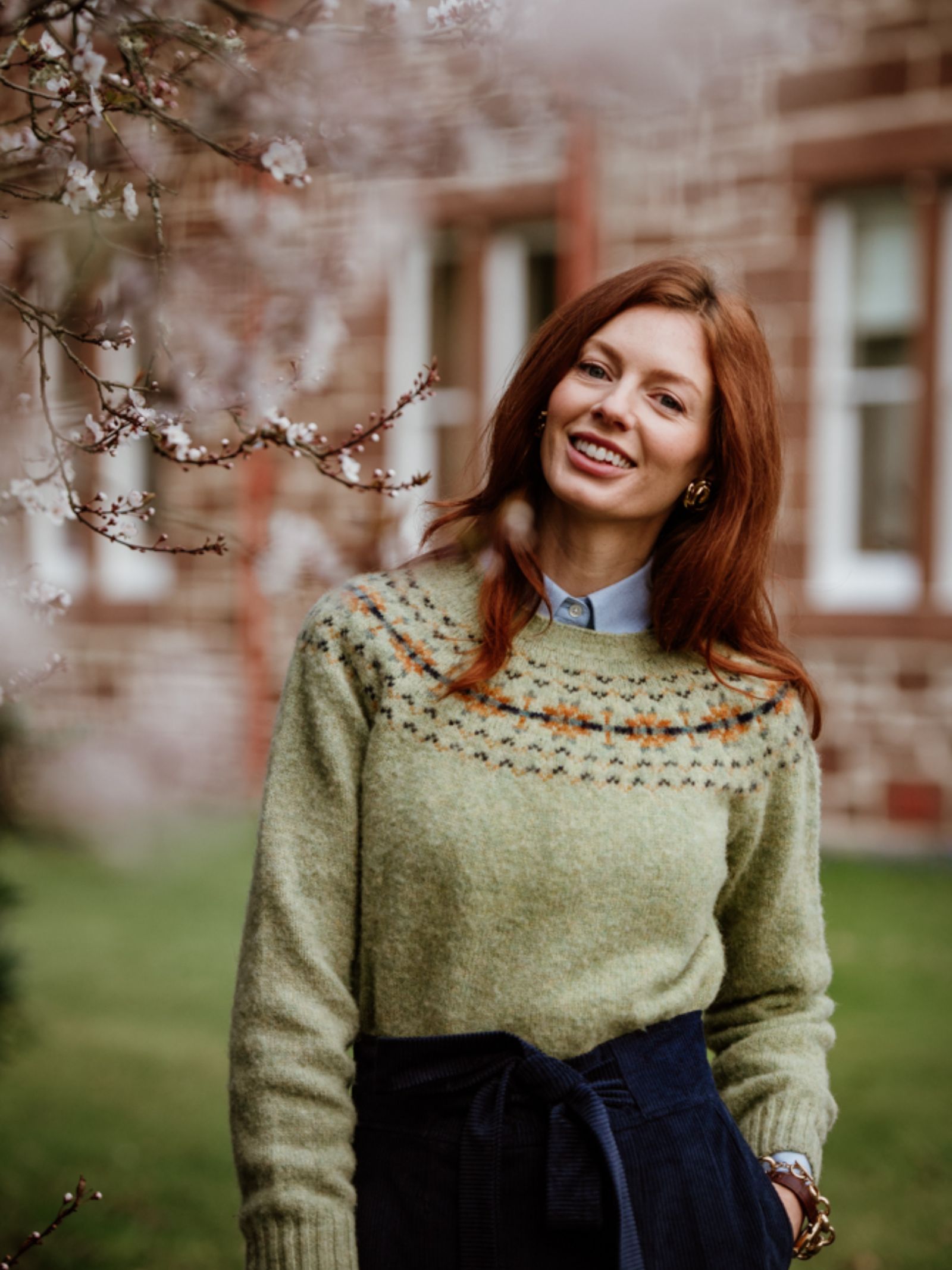 A woman with long red hair smiles outdoors by blooming branches, wearing the Campbell's of Beauly Auld Stock Cropped Fairisle Crew Neck Jumper and a dark skirt. A stone building with windows is in the background.