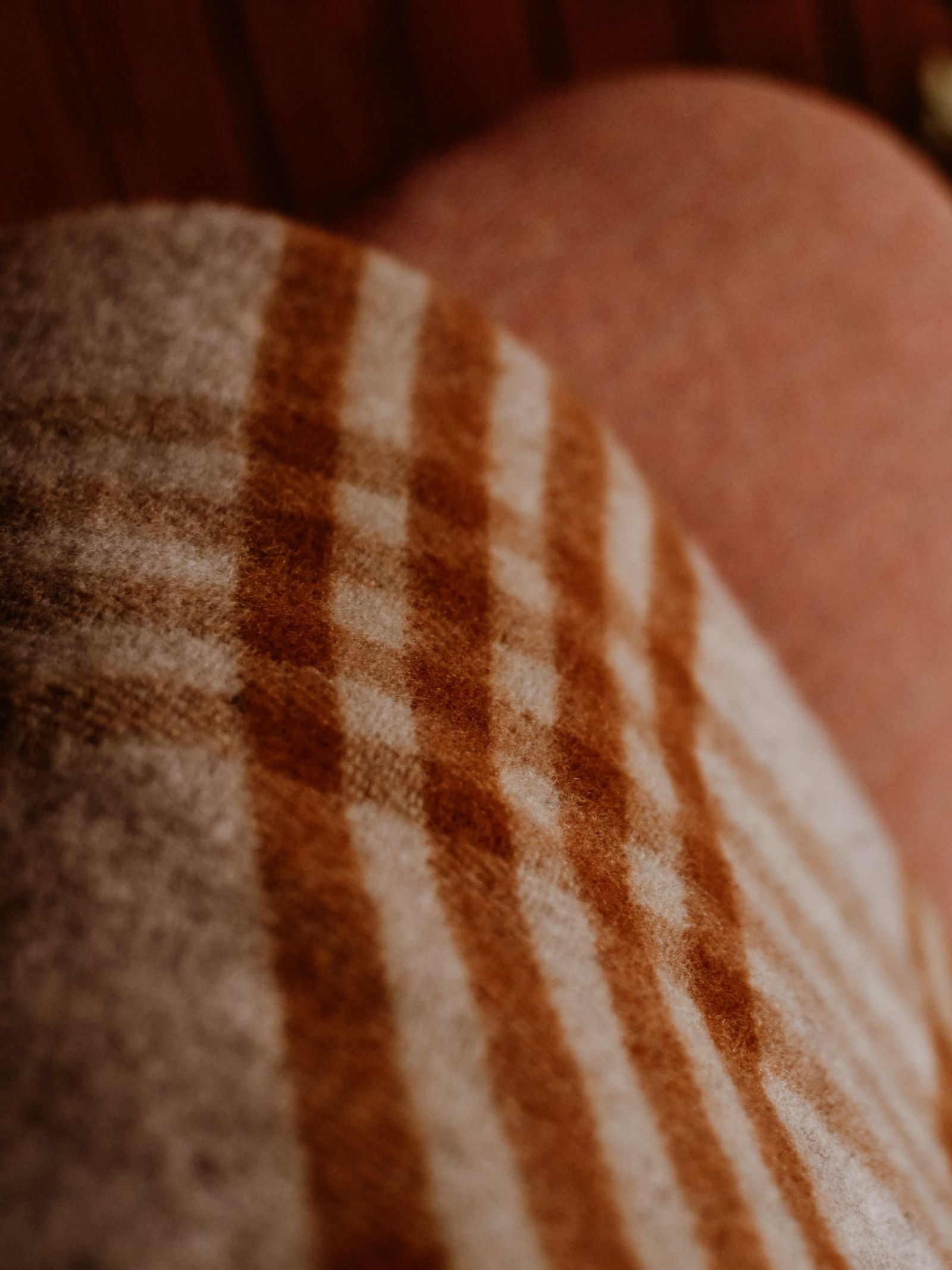 A Tarradale Lambswool Throw by Campbell's of Beauly, featuring a brown and cream check design, is draped over a person's leg beneath warm lighting, highlighting its soft texture and cozy atmosphere.