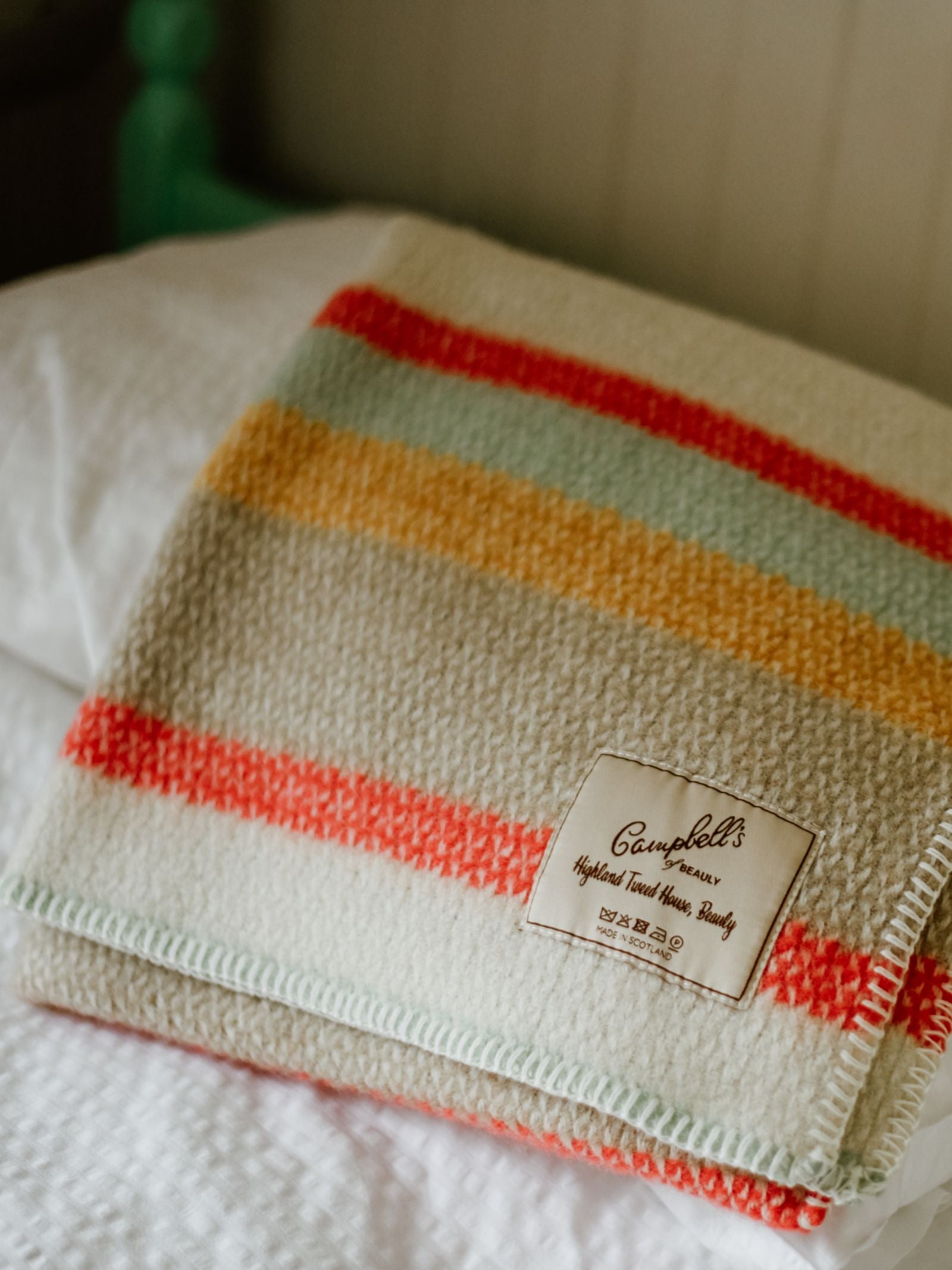 A folded Baby Blanket by Campbell's of Beauly, featuring red, yellow, and green stripes, rests on a white quilted bed. A sewn label displays the brand name and "Highest Trad House Quality.