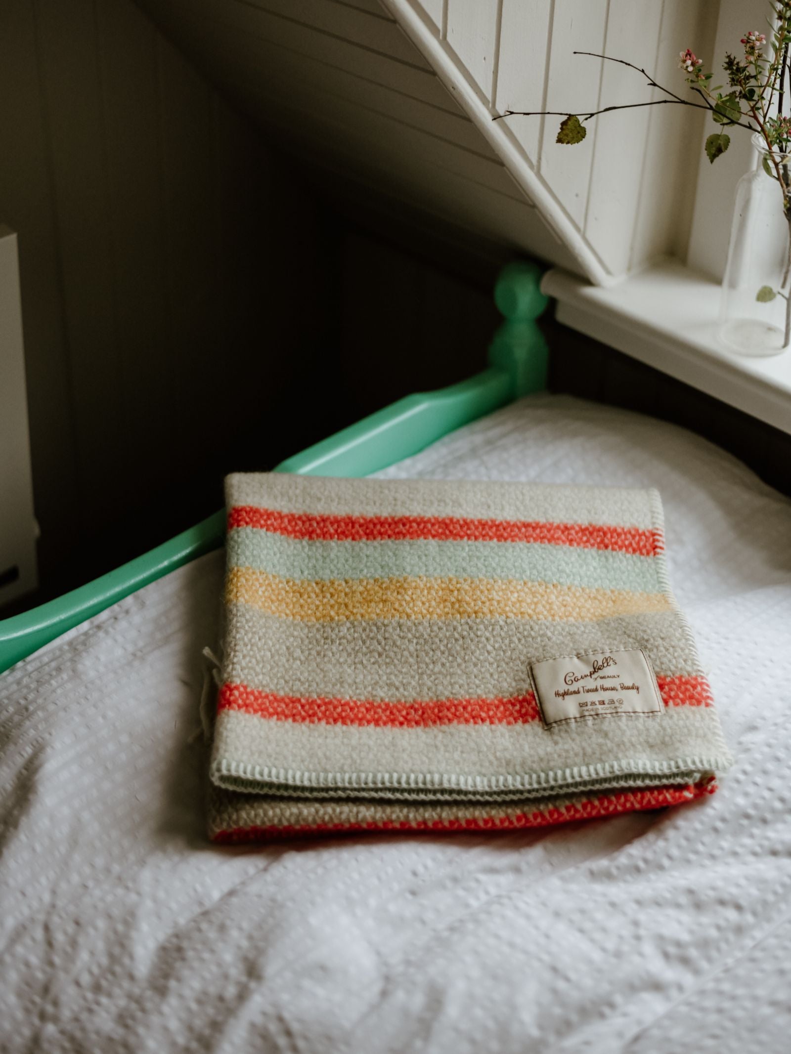 A Campbell's of Beauly Baby Blanket, striped in red, yellow, and beige wool, lies folded on a white bed with a green frame. A nearby window showcases a small vase of flowers.